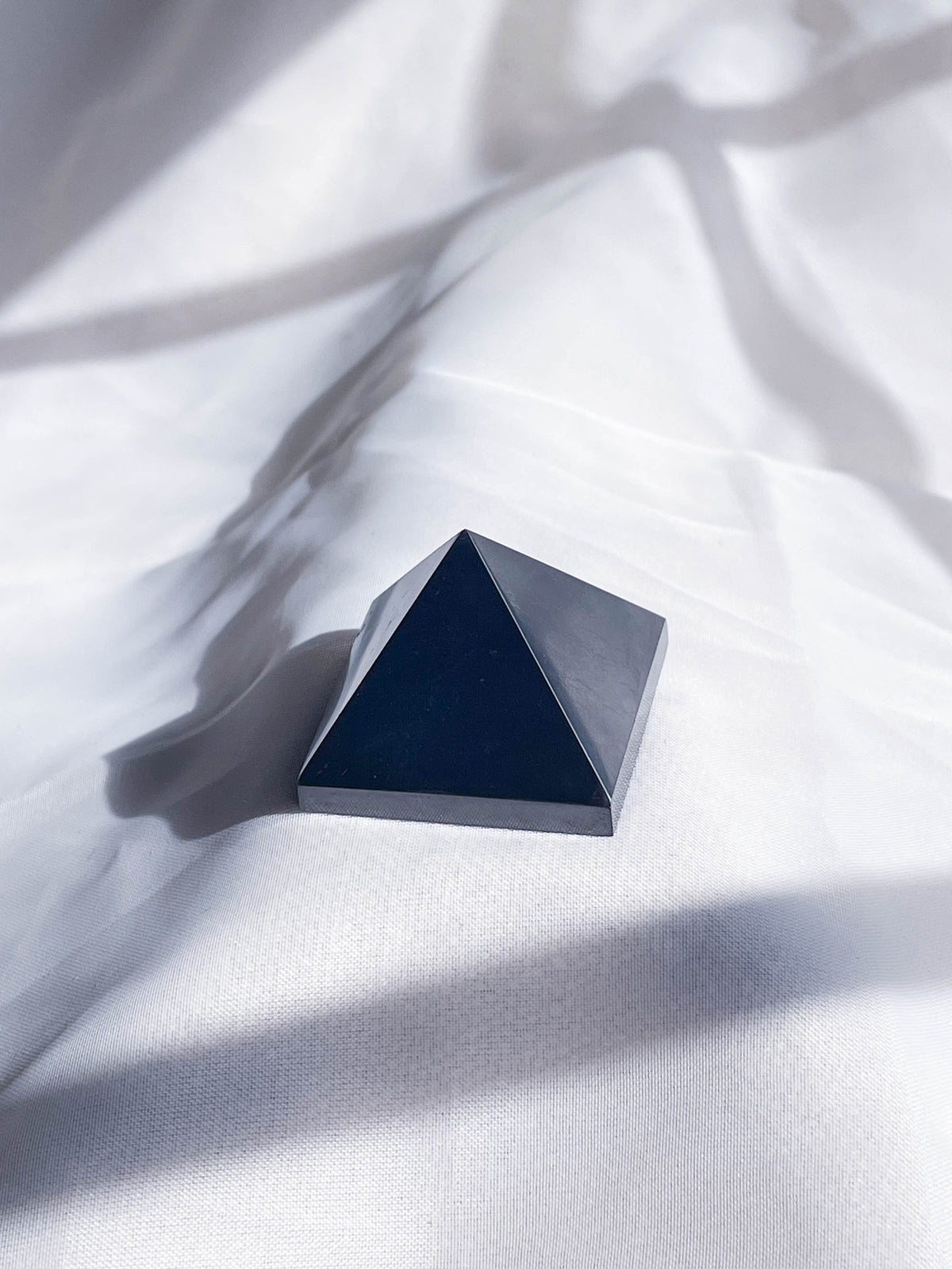 The Healing Hedge Witch's Shunghite Pyramid Point, intuitively chosen, rests on softly wrinkled white fabric as natural light and shadows play across the scene.