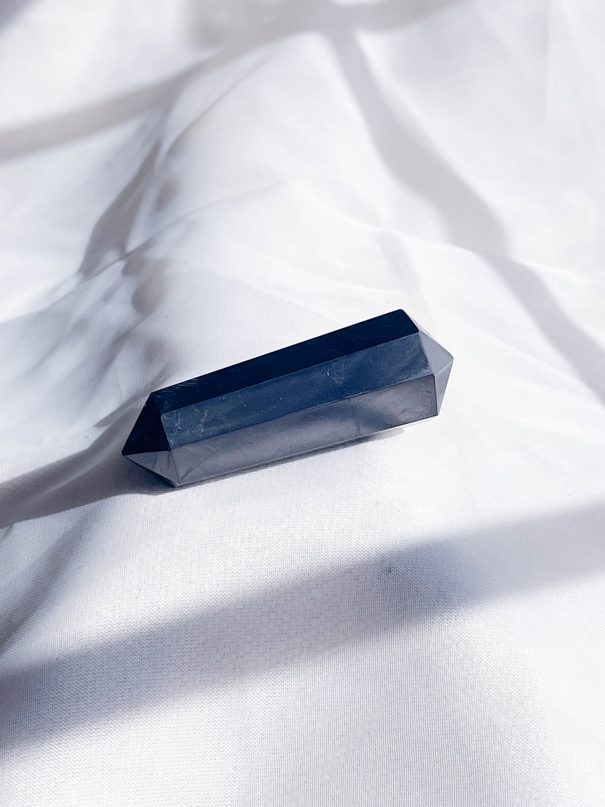 The Healing Hedge Witch's Shunghite Double Terminated Mini Crystal Tower rests on softly wrinkled white fabric, casting gentle shadows in natural light.