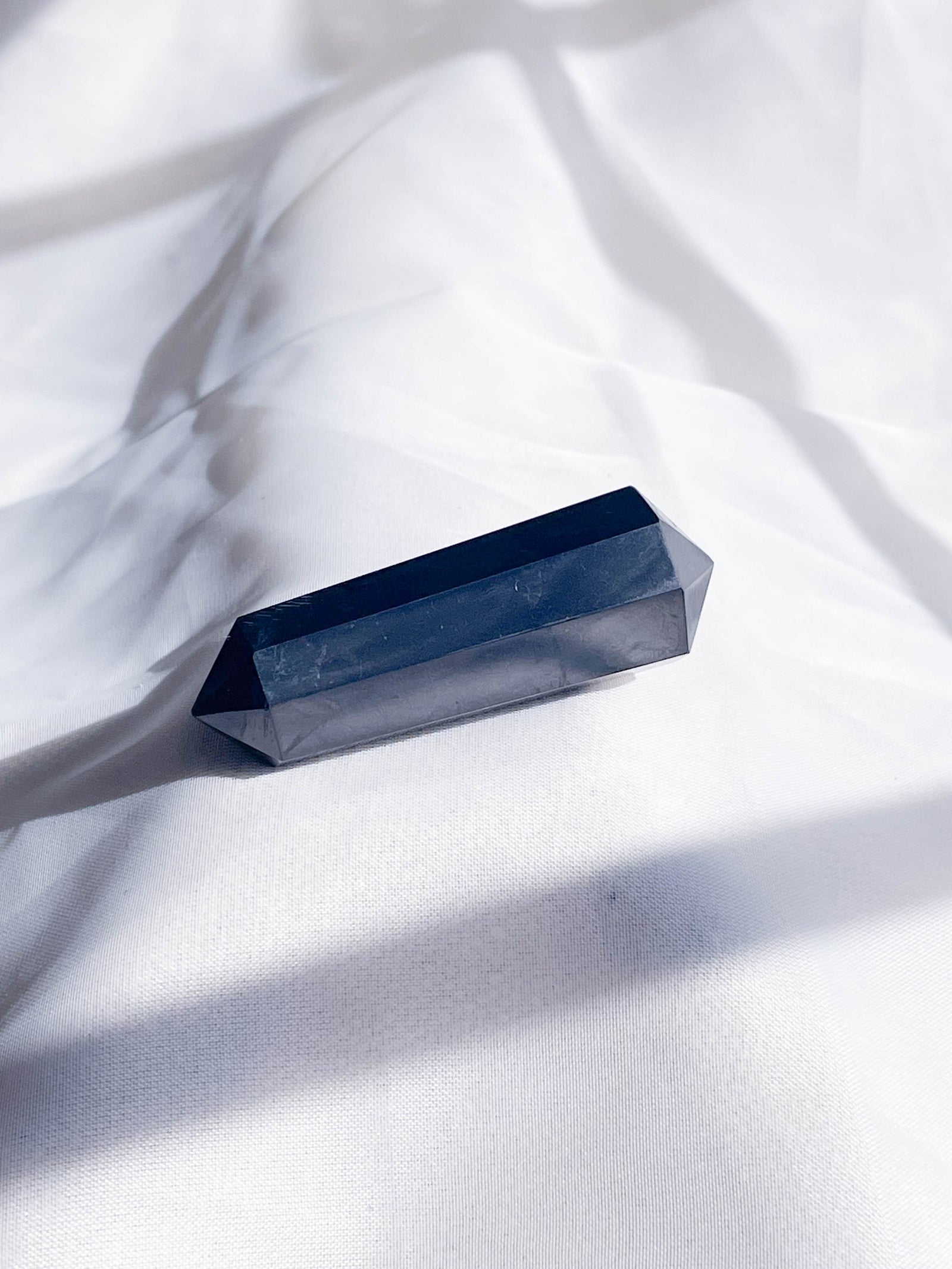 The Healing Hedge Witch's Shunghite Double Terminated Mini Crystal Tower rests on softly wrinkled white fabric, casting gentle shadows in natural light.