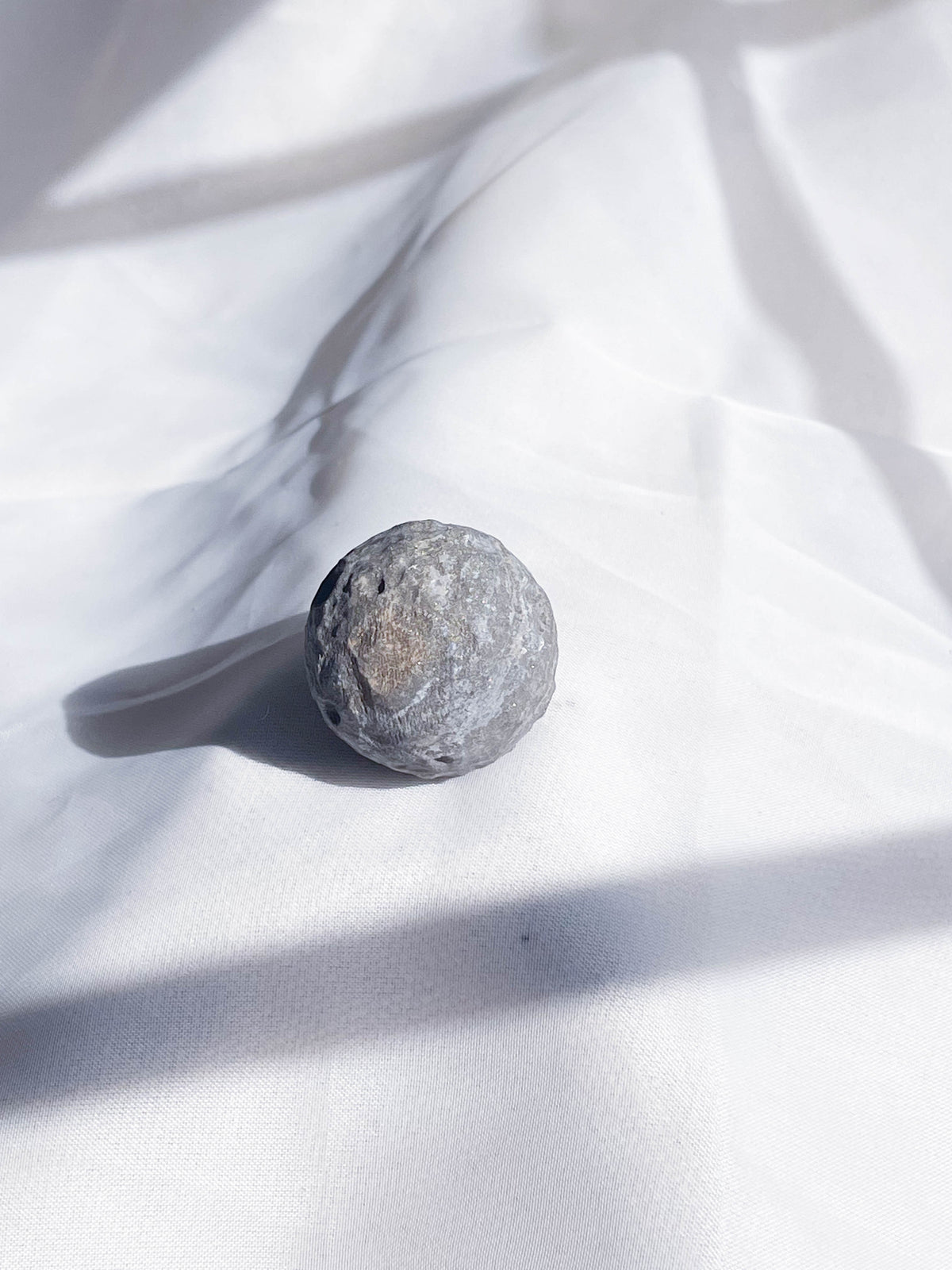 The Healing Hedge Witch’s Crater Crystal Moon, a small, round gray stone, rests on softly wrinkled white fabric as sunlight and shadows play across its surface.