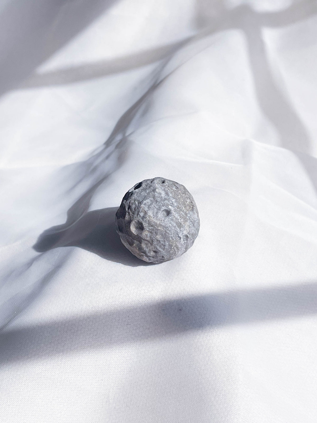 The Healing Hedge Witch's Crater Crystal Moon, a small, round gray stone with a rough, cratered surface, rests on softly lit white fabric with gentle shadows.