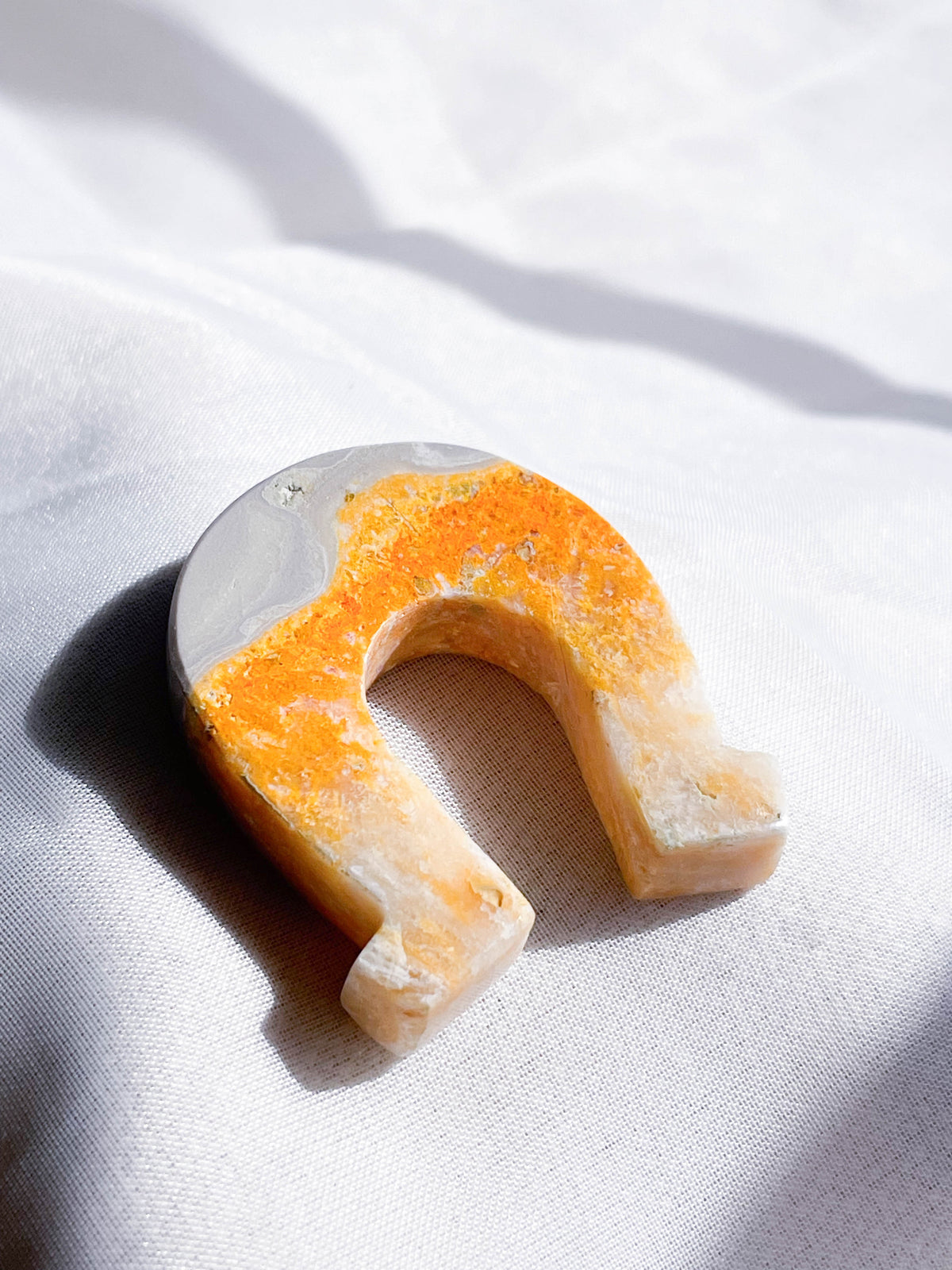 The Healing Hedge Witch's Bumblebee Bee Jasper Shape, a horseshoe-shaped stone in orange, yellow, and gray, rests on soft white fabric under bright natural light.