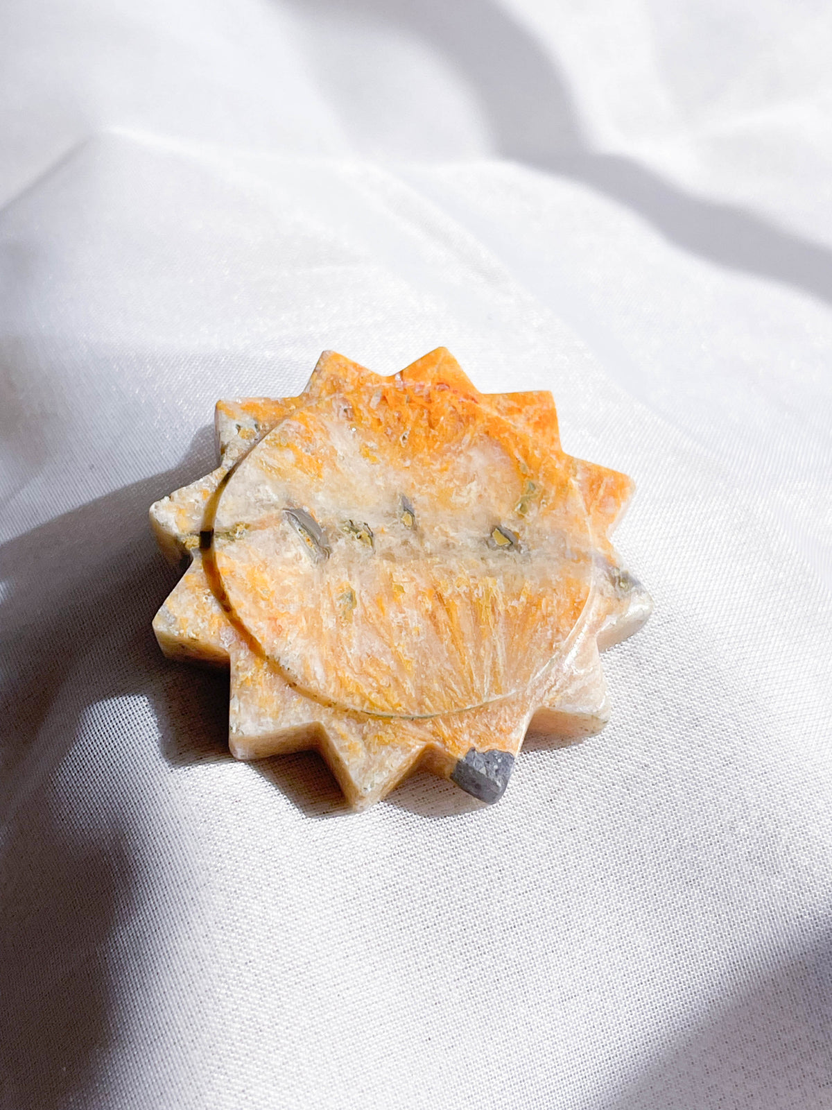 The Healing Hedge Witch's Bumblebee Bee Jasper Shape, an orange and white sun-shaped stone with subtle veins, rests on soft white fabric. Bright sunlight accentuates its marbled texture and unique coloration.