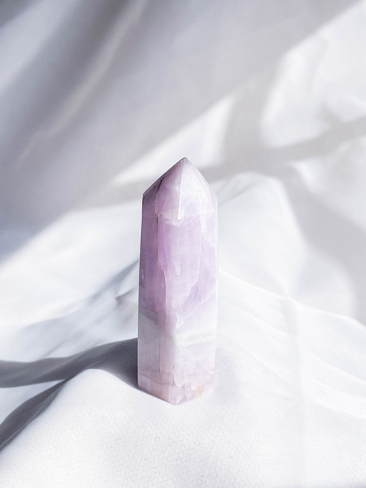 A Kunzite Mini Crystal Tower by The Healing Hedge Witch stands upright on soft white fabric, its polished light purple point illuminated by natural light and gentle shadows.