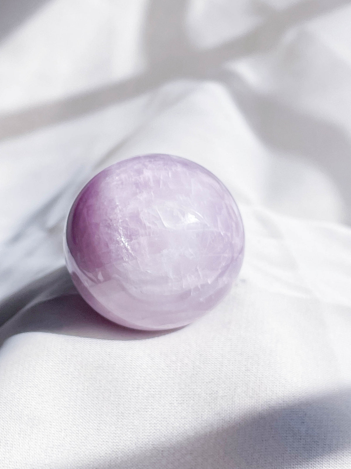 A smooth, polished Kunzite Sphere from The Healing Hedge Witch sits on soft white fabric, its light purple surface gently illuminated by natural light.