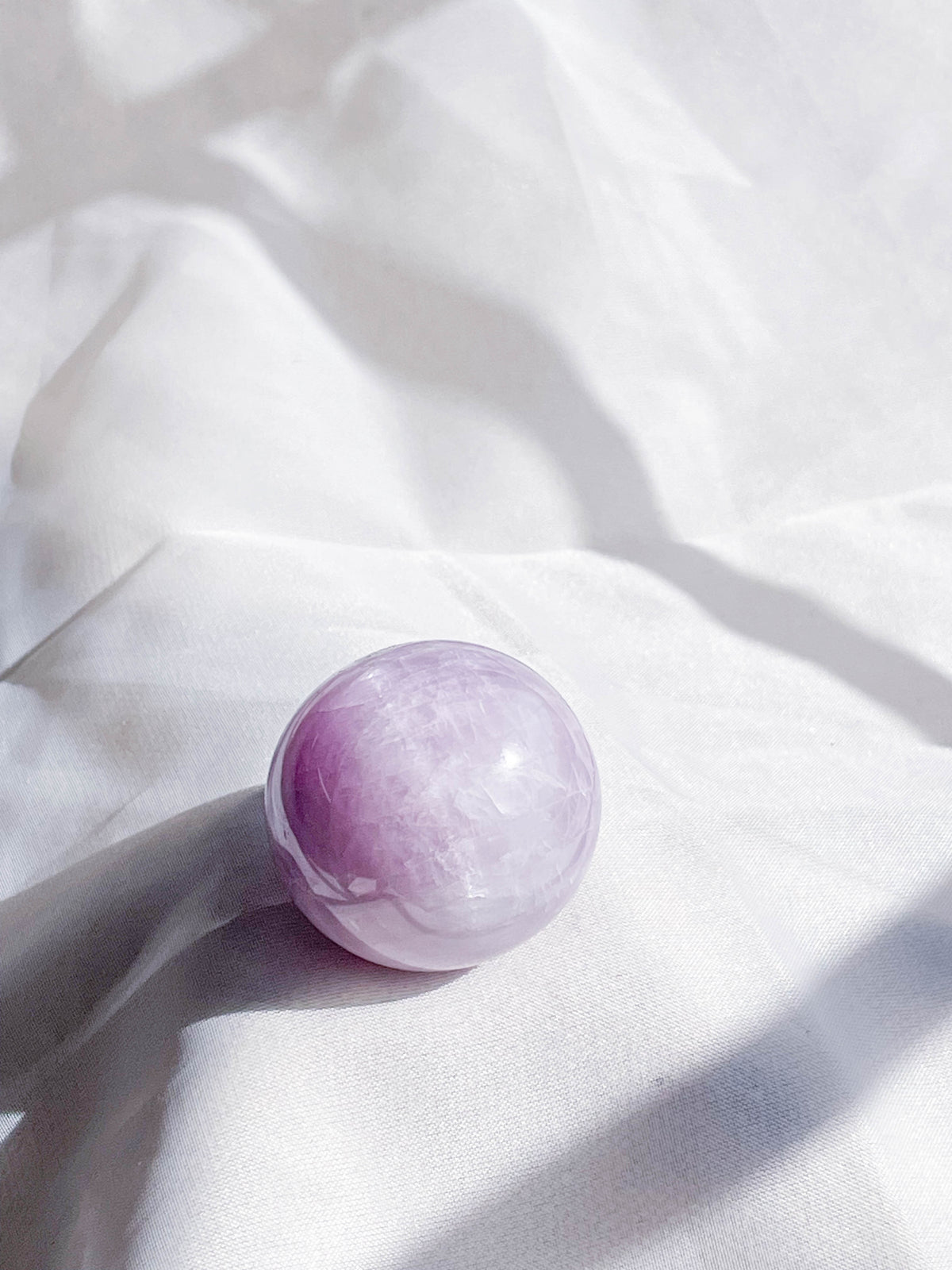 A polished Kunzite Sphere by The Healing Hedge Witch, in a light purple hue, rests on soft white fabric as gentle sunlight and shadows play across the scene.