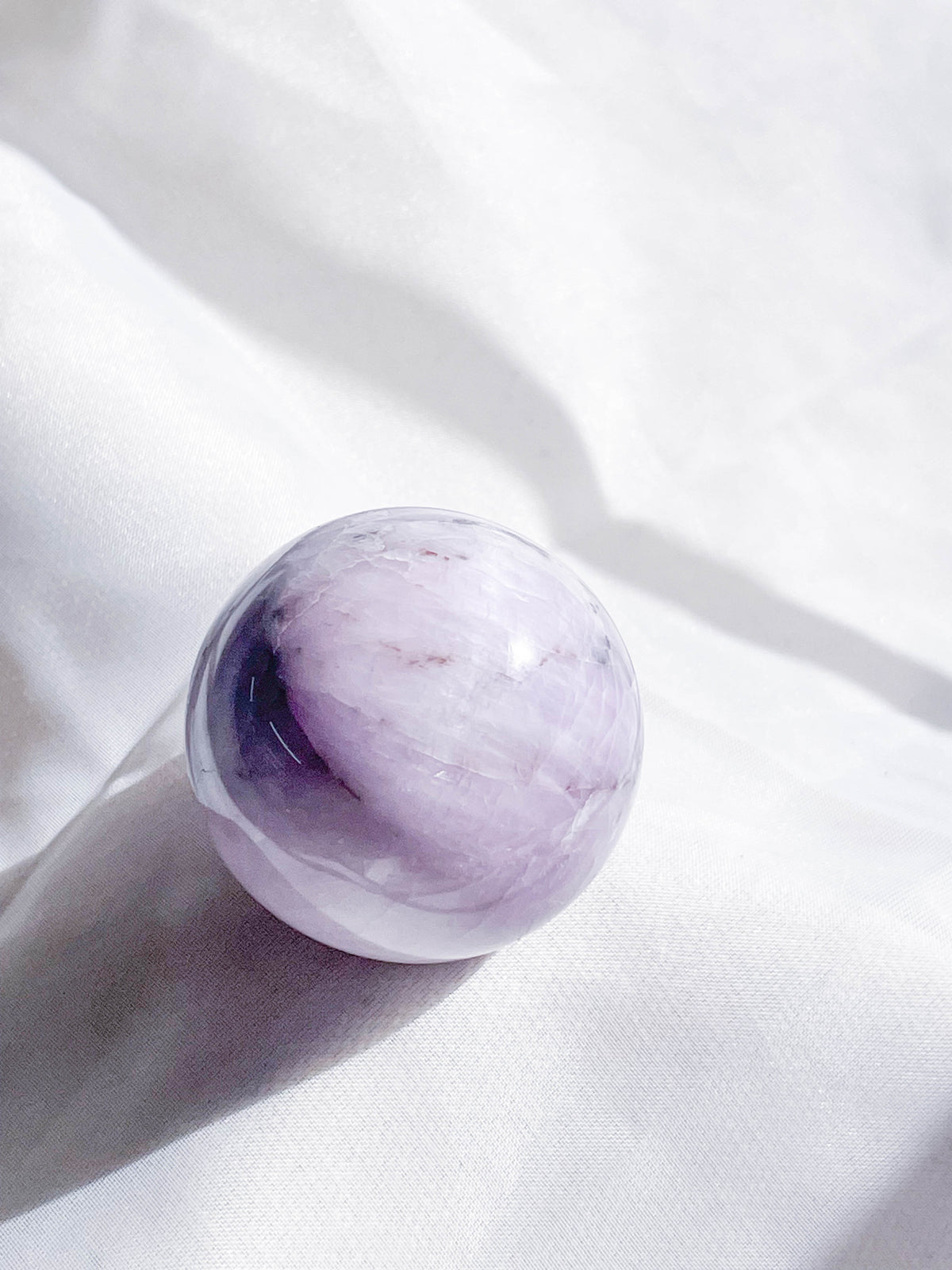 The Healing Hedge Witch Kunzite Sphere, a polished, round lavender and white crystal sphere, rests on soft white fabric and is illuminated by natural light.