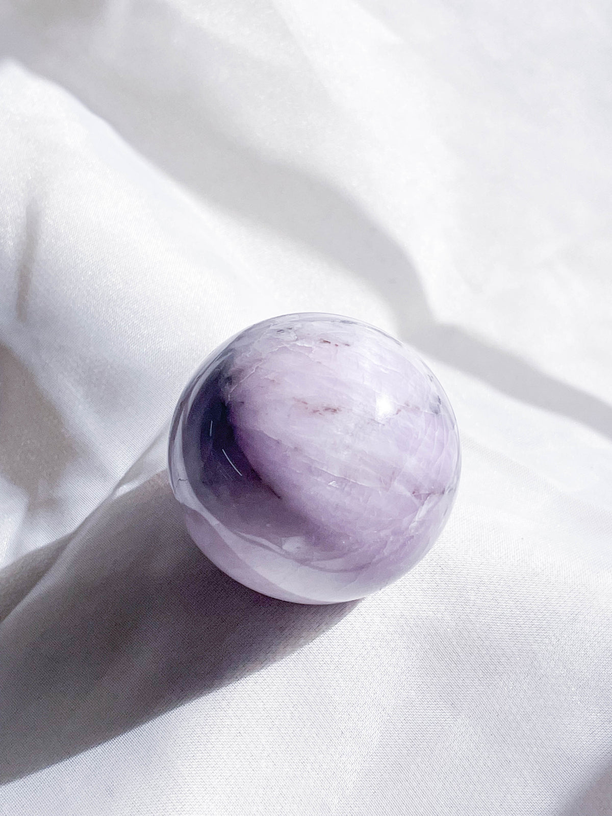 The Healing Hedge Witch Kunzite Sphere, a smooth, round light purple crystal, rests on softly wrinkled white fabric, casting a gentle shadow in bright natural light.