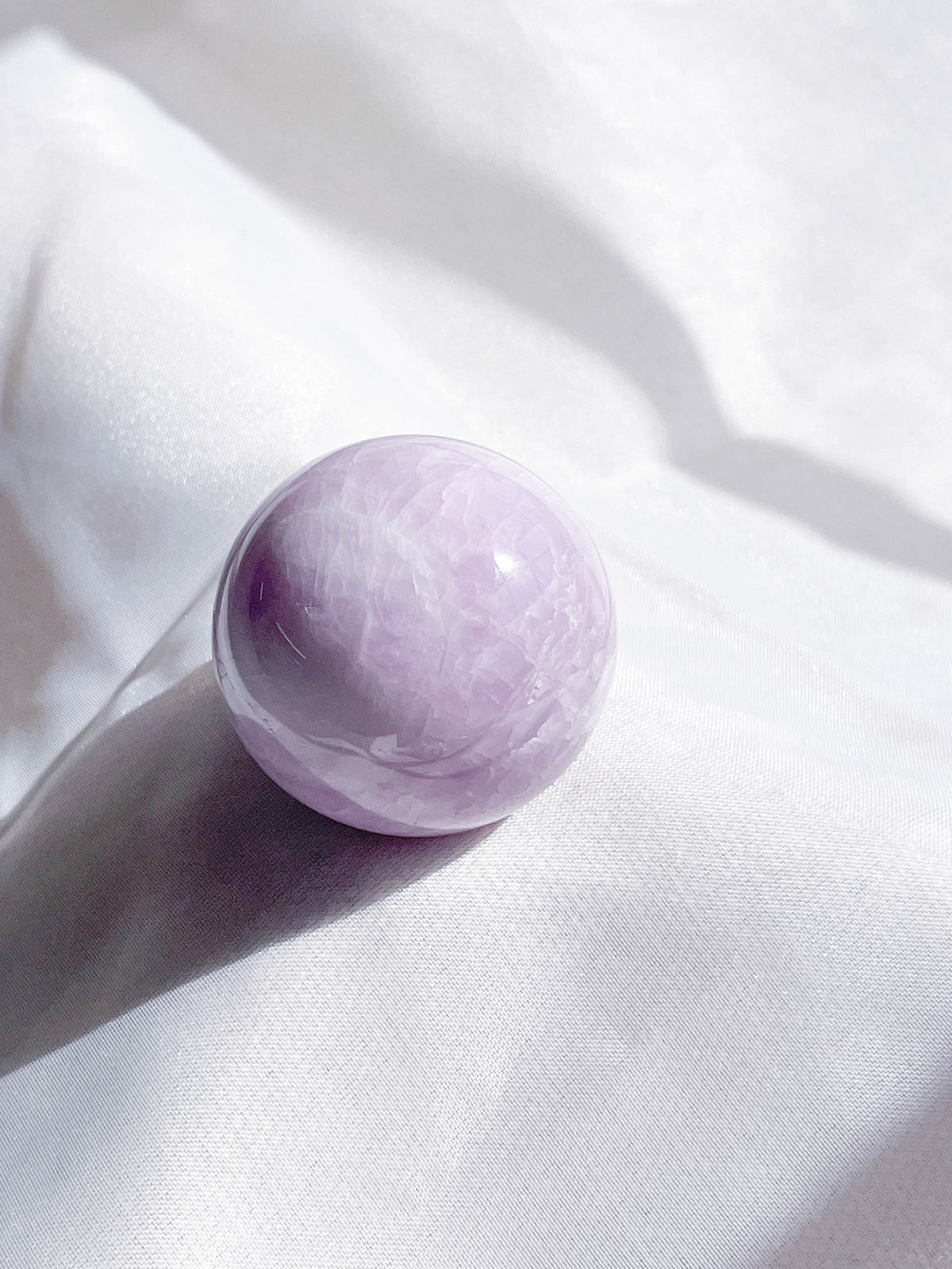 The Healing Hedge Witch Kunzite Sphere, a smooth, round, light purple crystal, rests on soft white fabric and is illuminated by natural light that reveals gentle shadows and highlights.