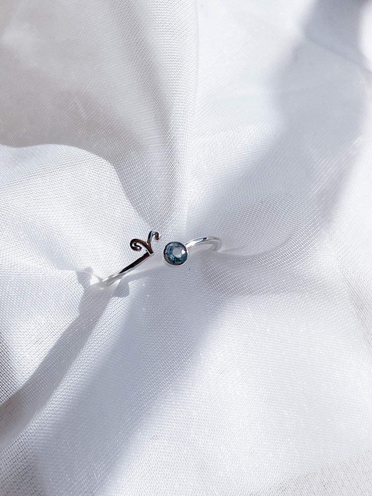 The Healing Hedge Witch’s Zodiac Sterling Silver Ring, featuring the Aries symbol and a small blue birthstone, is displayed on soft white fabric.