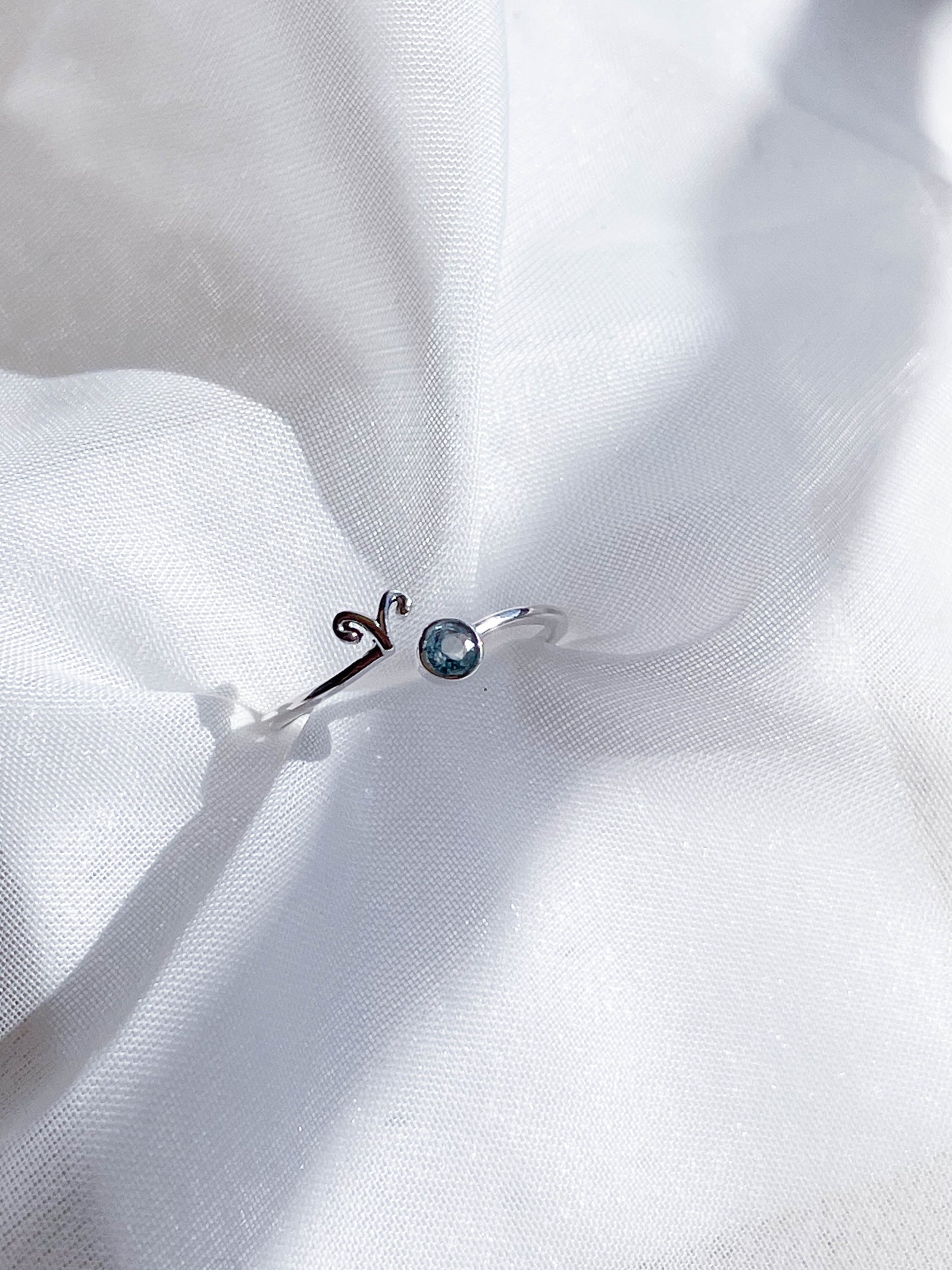 The Healing Hedge Witch’s Zodiac Sterling Silver Ring, featuring the Aries symbol and a small blue birthstone, is displayed on soft white fabric.