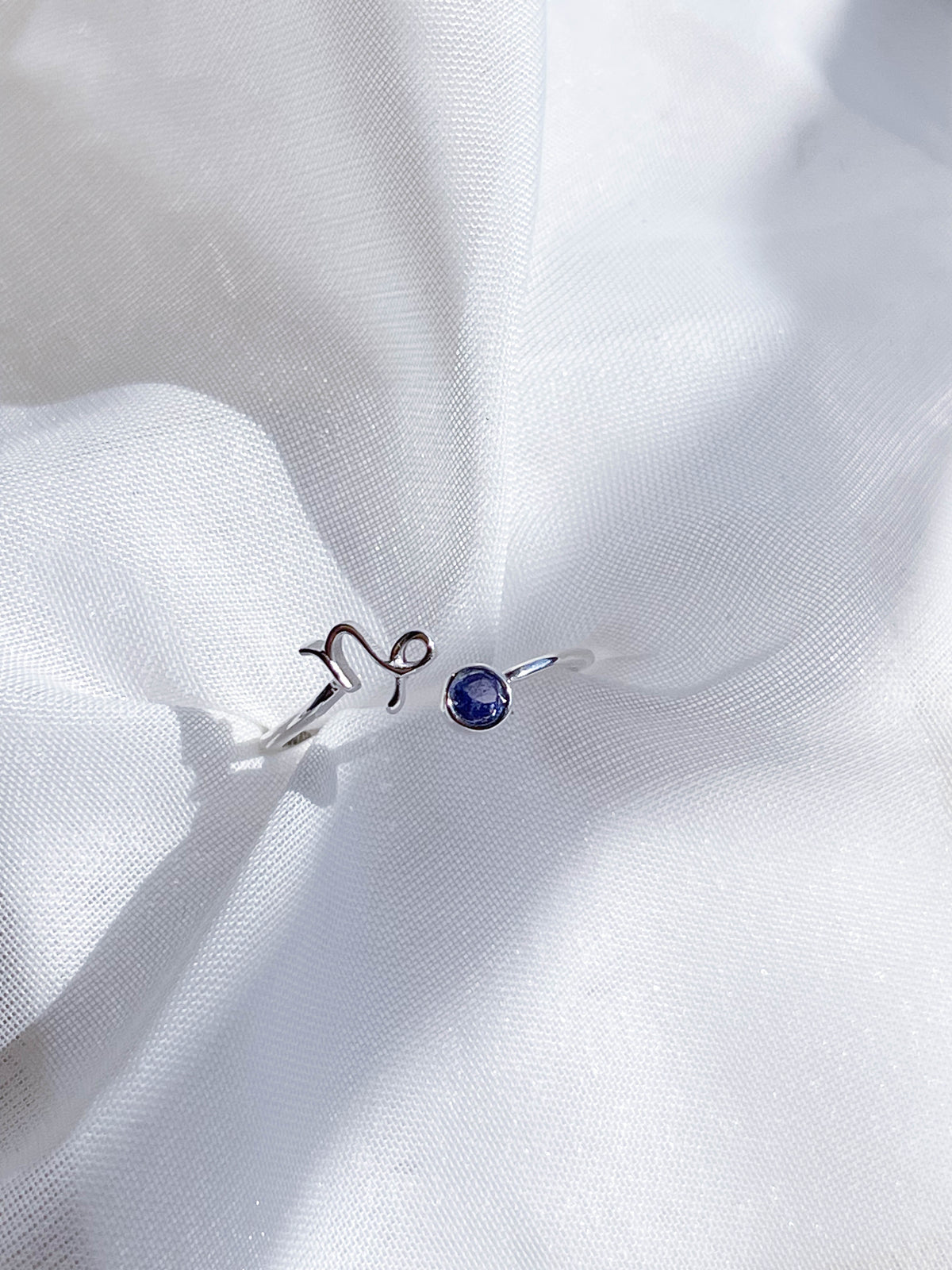 The Healing Hedge Witch Zodiac Sterling Silver Ring, adorned with a minimalist Leo symbol and small blue birthstone, is showcased on softly textured white fabric.
