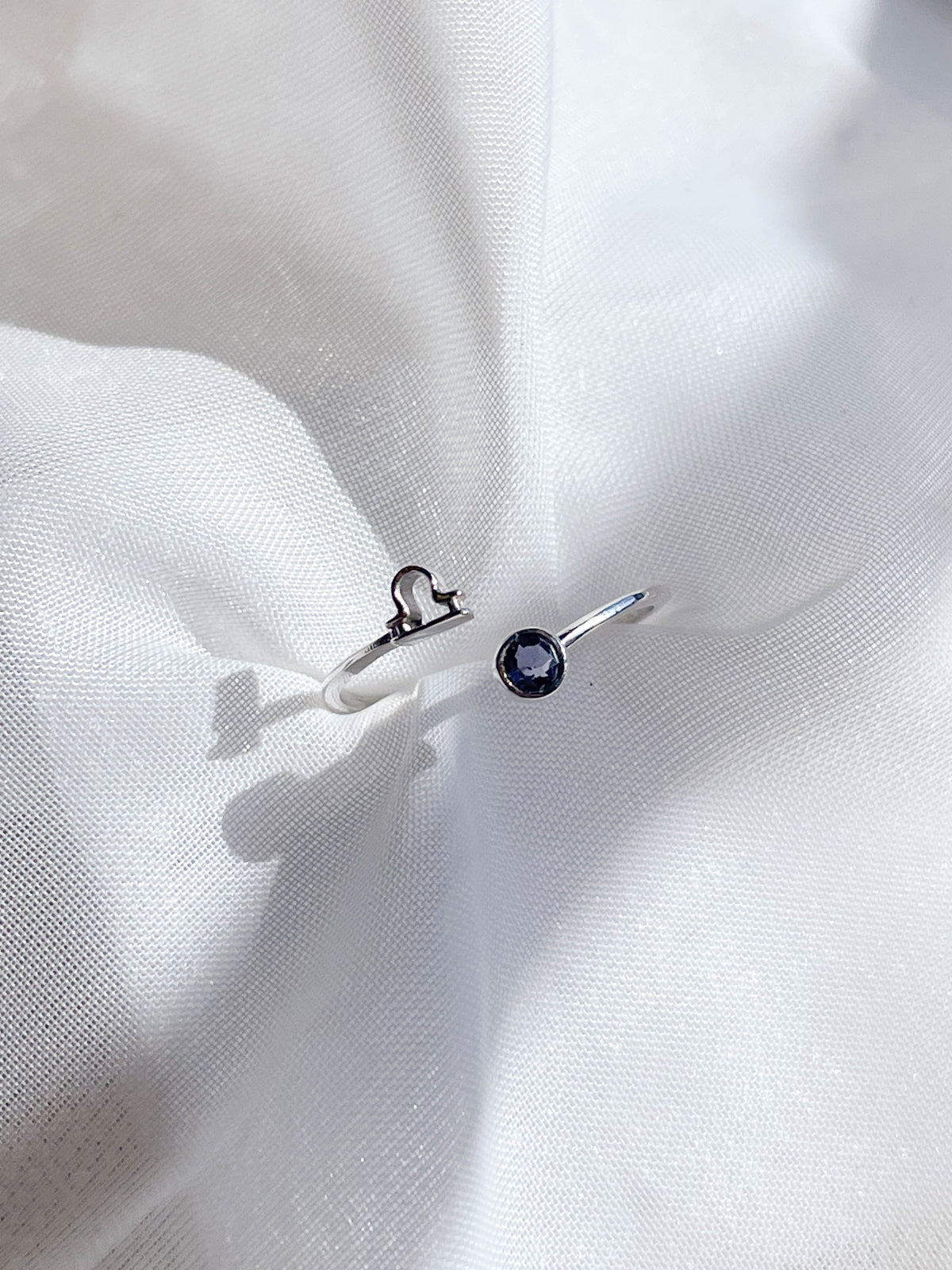 The Healing Hedge Witch’s Zodiac Sterling Silver Ring features an open design with a dark round birthstone on one end and a small Libra symbol on the other, shown on softly draped white fabric.