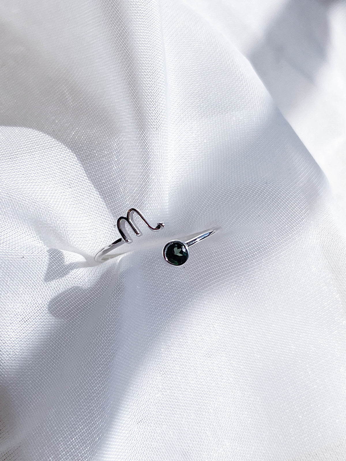 The Healing Hedge Witch Zodiac Sterling Silver Ring, with a minimalist Scorpio symbol and a small dark birthstone, is displayed on white wrinkled fabric in bright natural light.