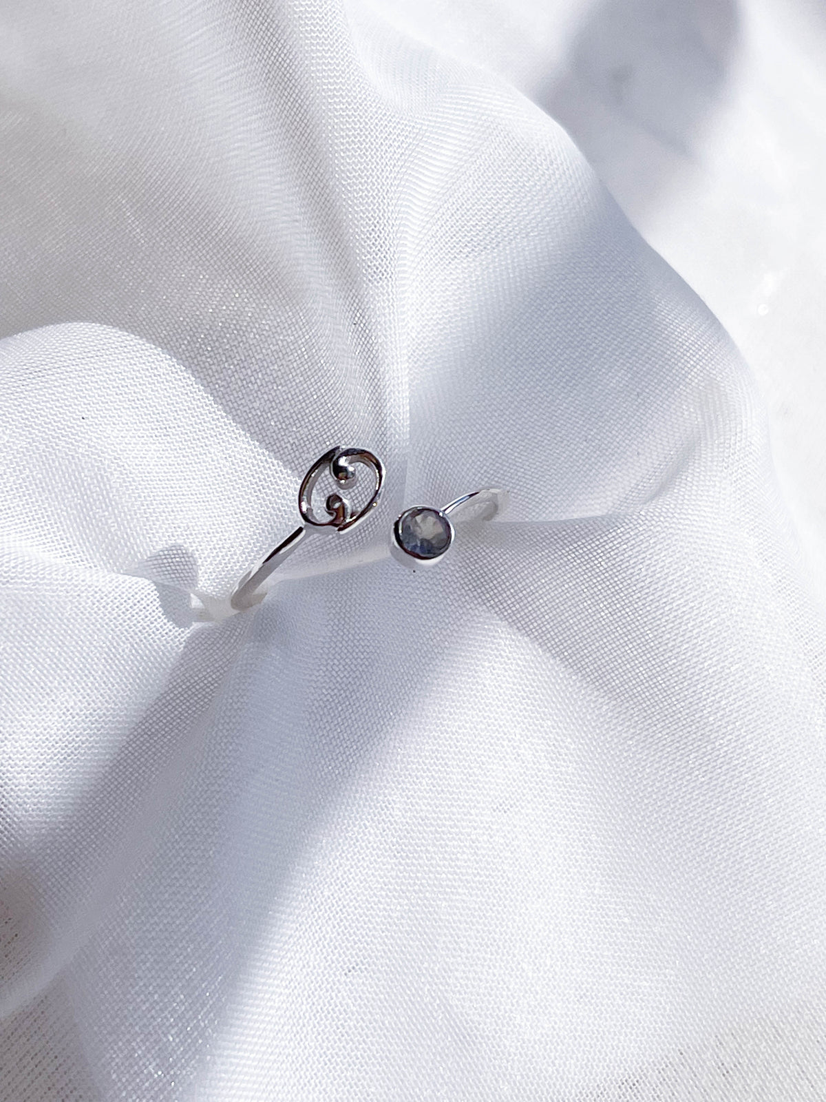The Healing Hedge Witch Zodiac Sterling Silver Ring features a round birthstone on one end and a small double G design on the other, shown resting on soft white fabric in bright natural light.