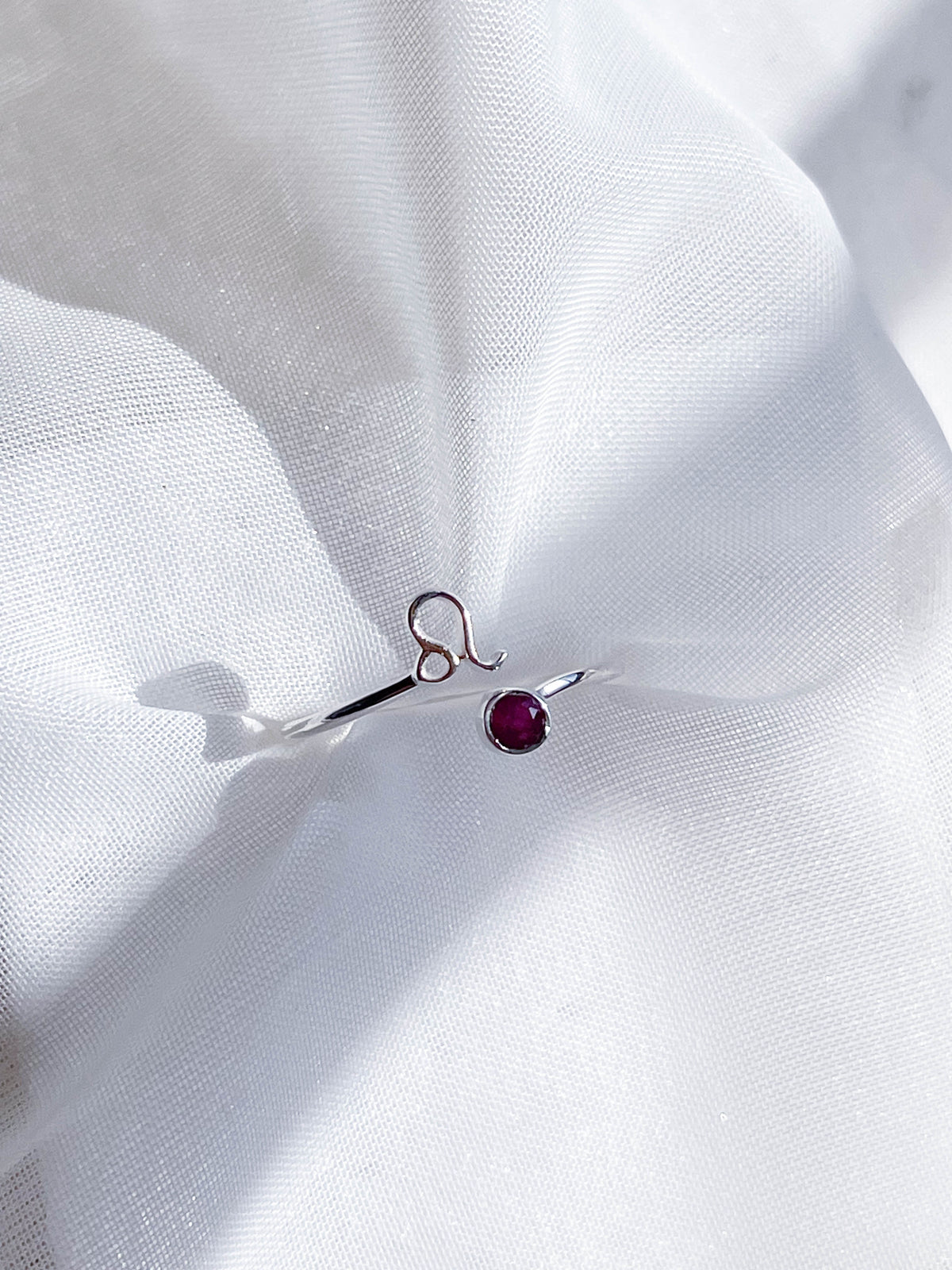 The Healing Hedge Witch’s Zodiac Sterling Silver Ring, featuring an abstract loop design and small red gemstone, rests on soft white textured fabric in bright natural light.