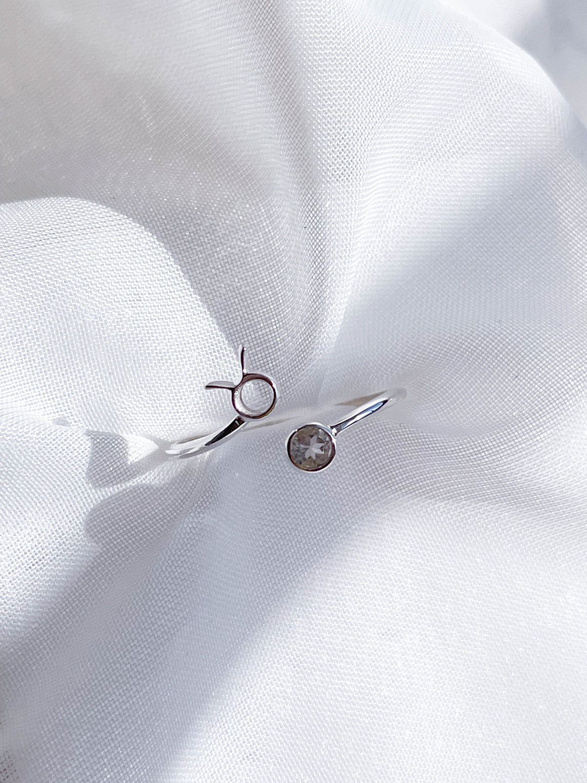 The Healing Hedge Witch's Zodiac Sterling Silver Ring features a minimalist open design with a small circular birthstone on one end and a simple metal circle on the other, displayed on softly textured white fabric.