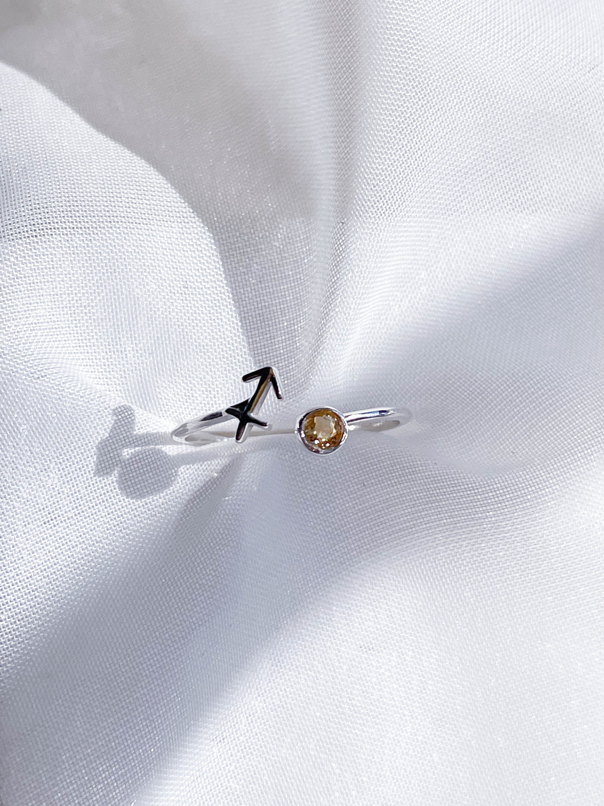 The Healing Hedge Witch's Zodiac Sterling Silver Ring features an arrow design on one end and a small yellow birthstone on the other, elegantly displayed on white textured fabric.