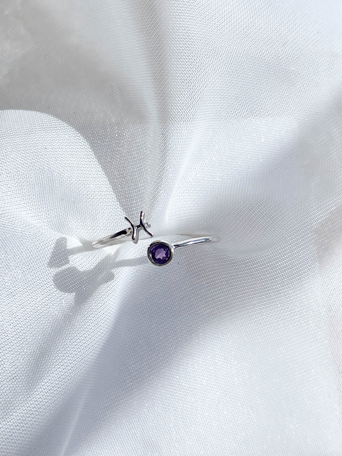 The Healing Hedge Witch’s Zodiac Sterling Silver Ring features an open design with a small purple birthstone on one end and a minimalist star on the other, shown resting on white textured fabric.