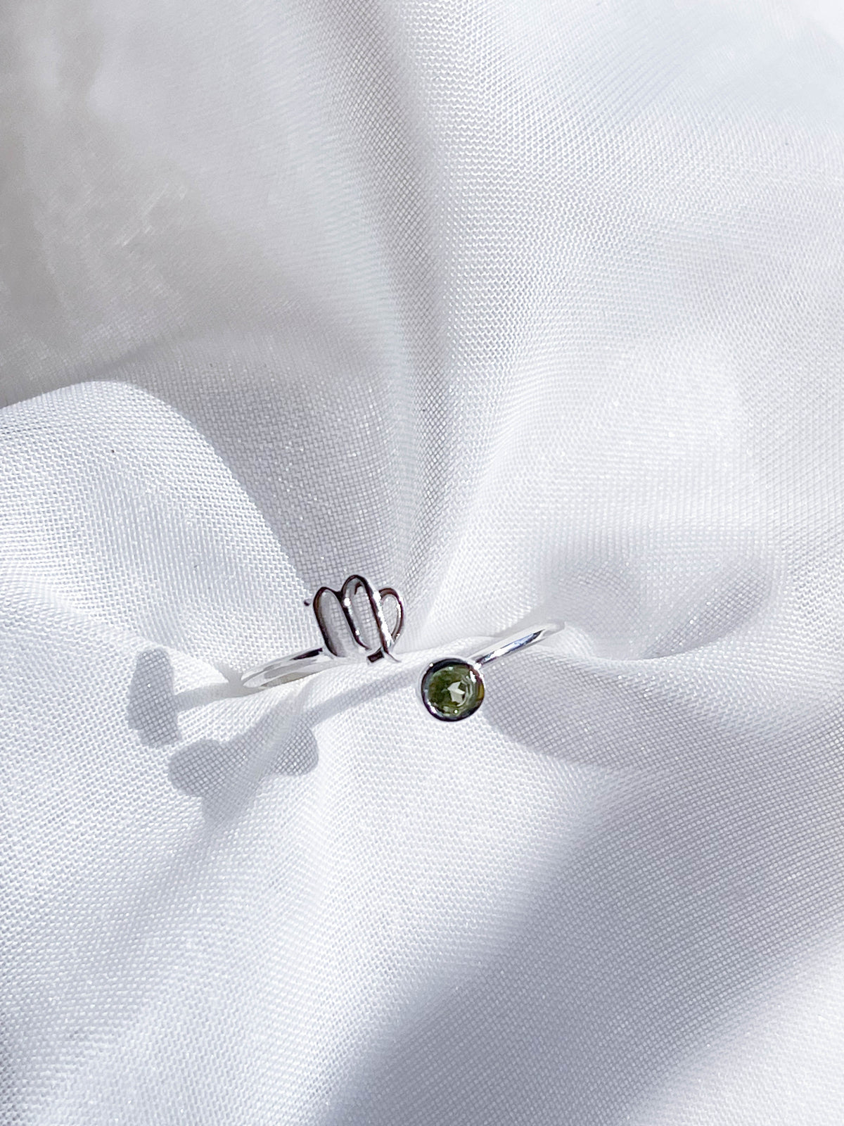 The Healing Hedge Witch's Zodiac Sterling Silver Ring, featuring a Virgo symbol and a green birthstone, is displayed on white textured fabric under soft light.