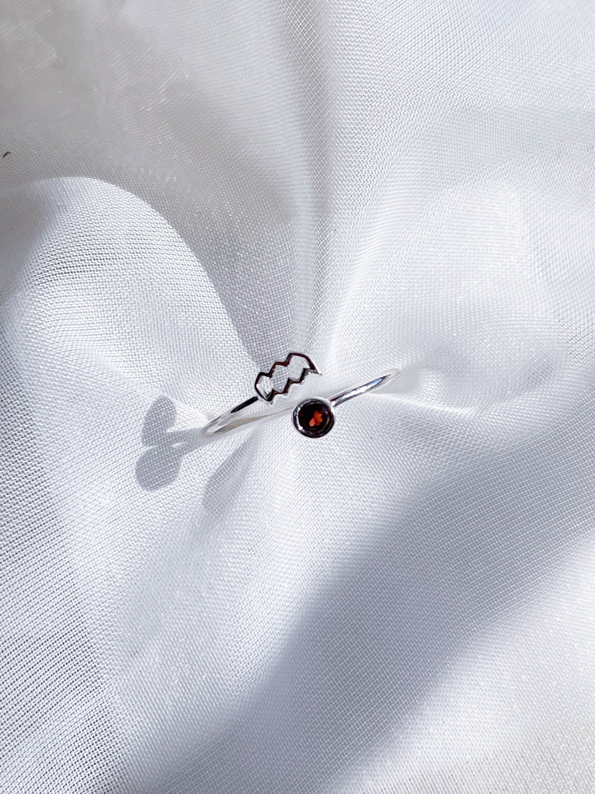 The Healing Hedge Witch’s Zodiac Sterling Silver Ring features a wavy Aquarius symbol and a small red birthstone, shown resting on white textured fabric—a stylish adjustable zodiac ring.