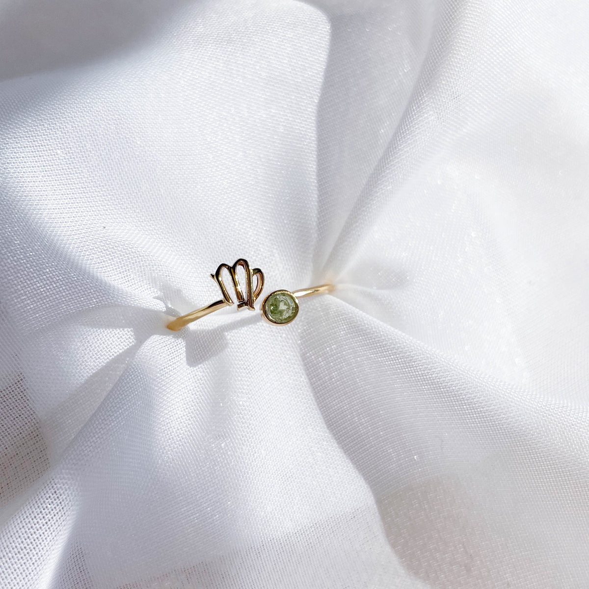 The Healing Hedge Witch's Zodiac Gold Plated Ring, featuring a Virgo symbol and small green birthstone, rests on soft, white, slightly crumpled fabric.