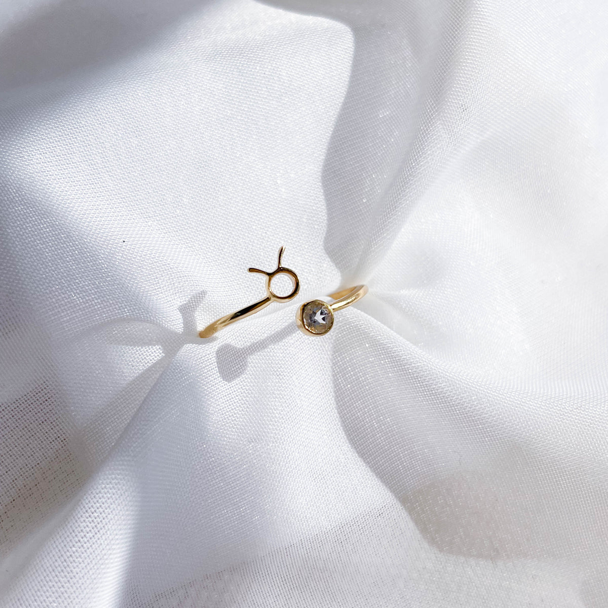 The Healing Hedge Witch Zodiac Gold Plated Ring, featuring a Taurus symbol and clear birthstone, gleams as it rests on softly wrinkled white fabric.