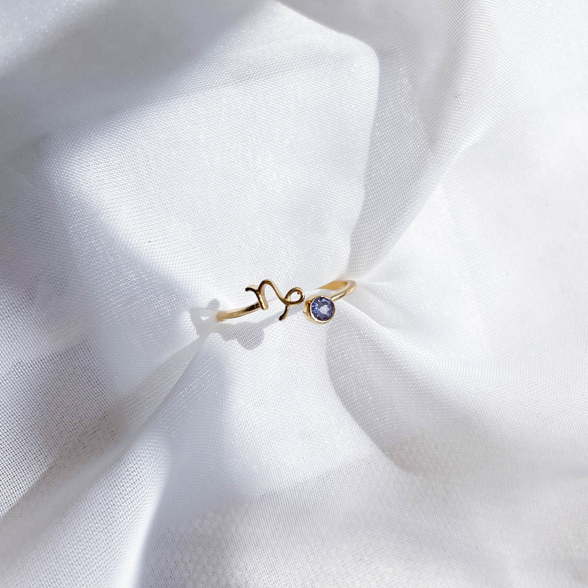 The Healing Hedge Witch's Zodiac Gold Plated Ring, featuring a small blue gemstone and wavy design, rests on softly wrinkled white fabric, glowing in gentle natural light.