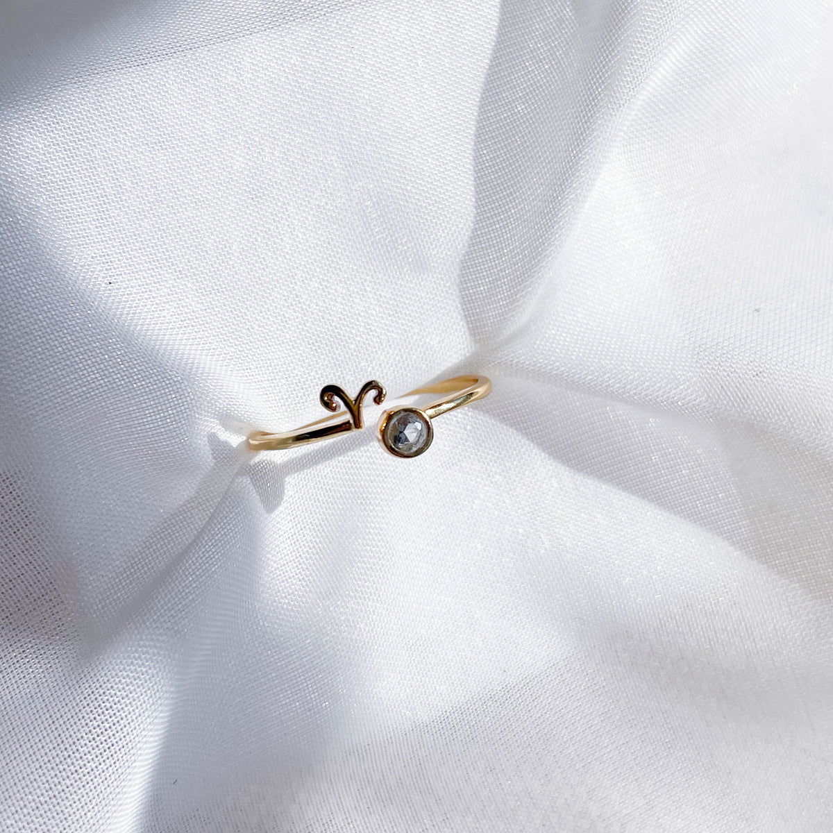 The Healing Hedge Witch Zodiac Gold Plated Ring, featuring the Aries symbol and a small round birthstone, sits on softly wrinkled white fabric in bright light.