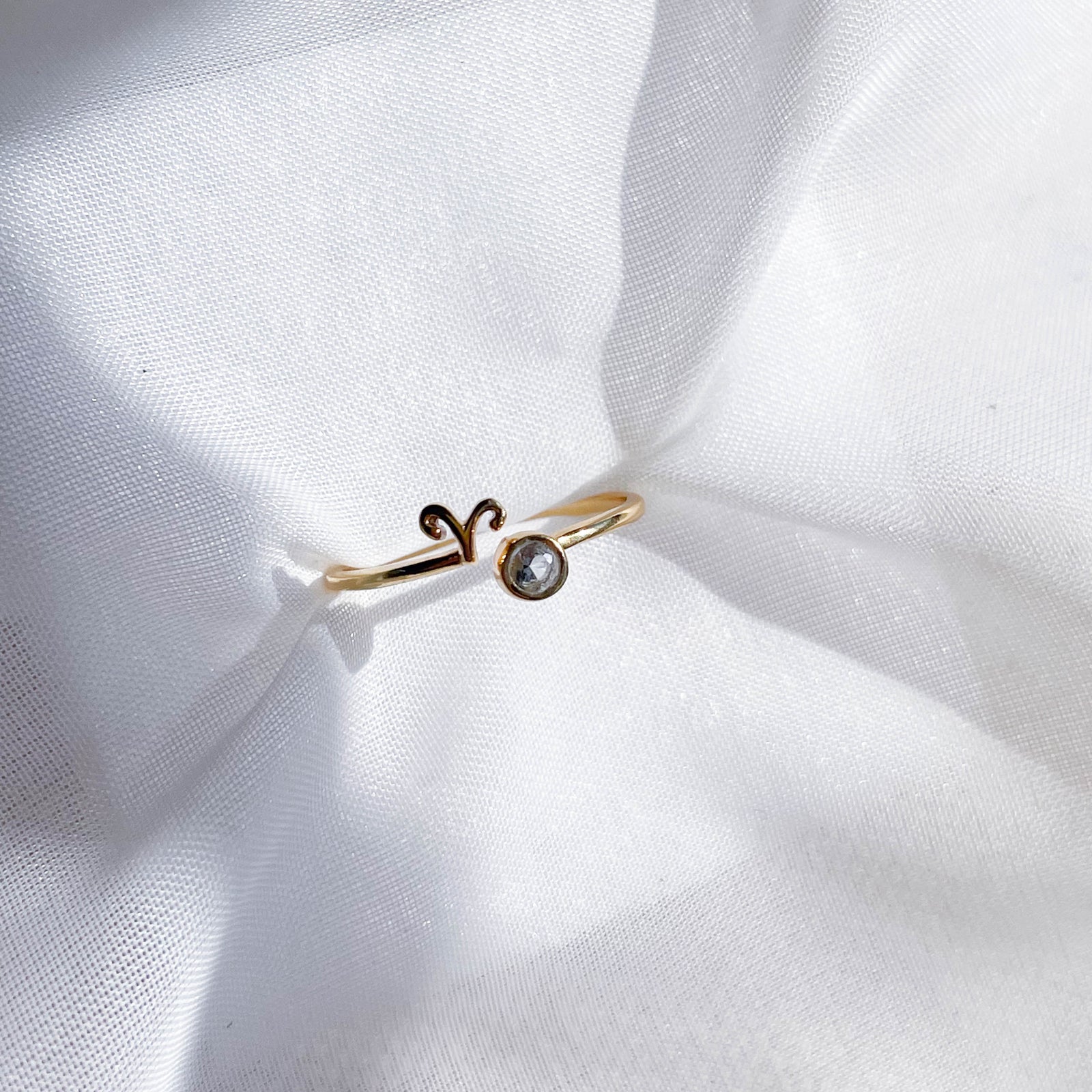 The Healing Hedge Witch Zodiac Gold Plated Ring, featuring the Aries symbol and a small round birthstone, sits on softly wrinkled white fabric in bright light.