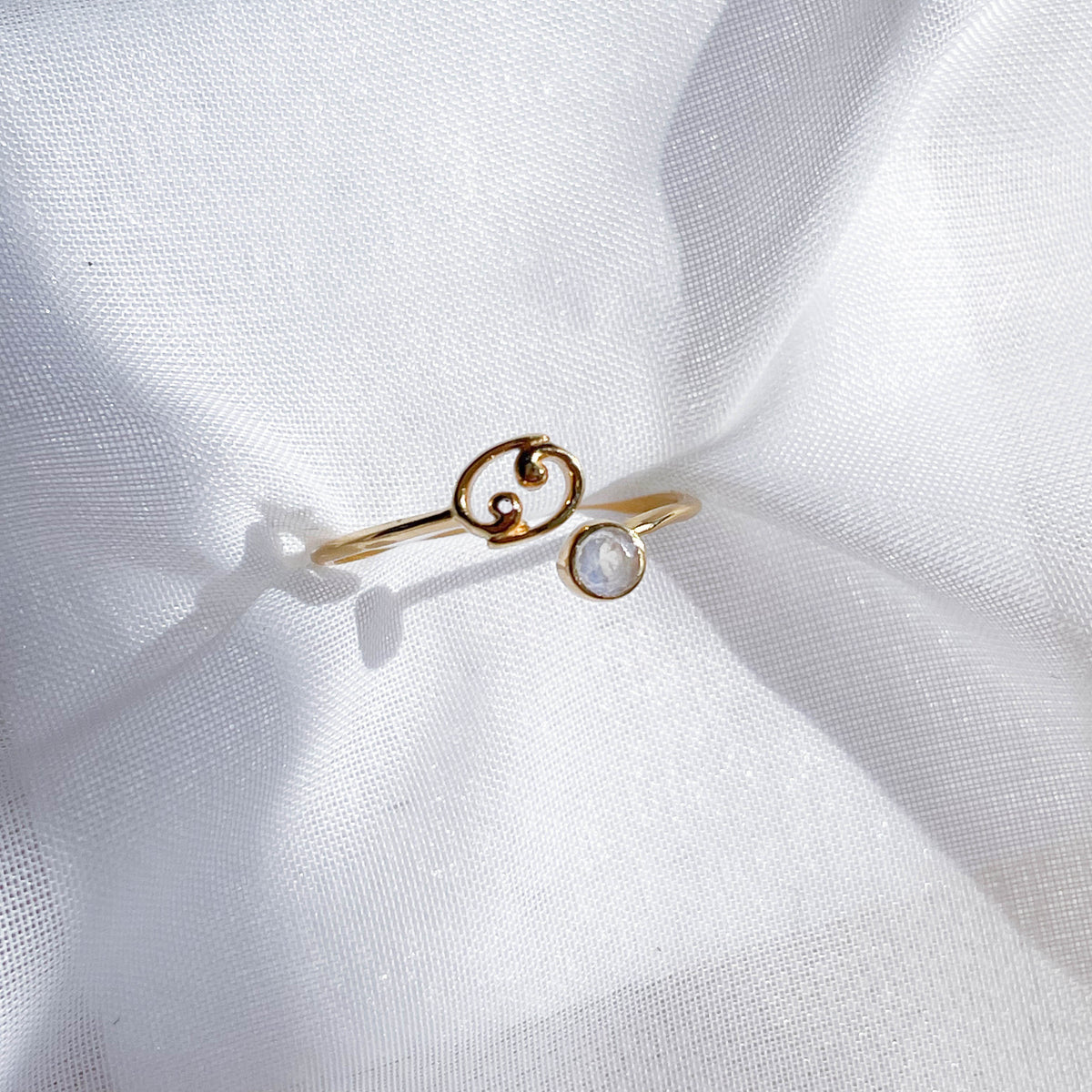 The Healing Hedge Witch’s Zodiac Gold Plated Ring features a swirl design and a single round white stone, shown resting on soft white fabric. This adjustable ring is perfect for any style.