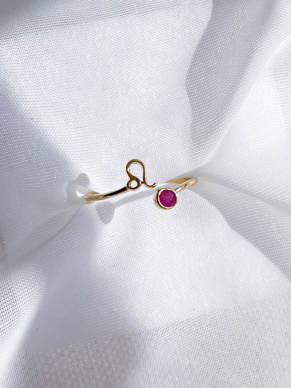 The Healing Hedge Witch Zodiac Gold Plated Ring features a Leo zodiac symbol and a small round pink birthstone, shown resting on gently folded white sheer fabric.