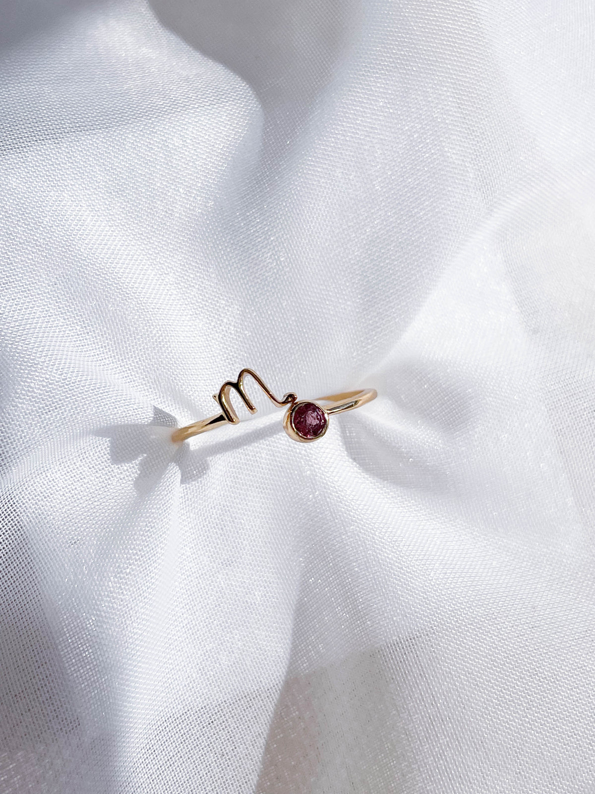 The Healing Hedge Witch’s Zodiac Gold Plated Ring, featuring a wavy design and a small red birthstone, is elegantly displayed on white sheer fabric under soft lighting.