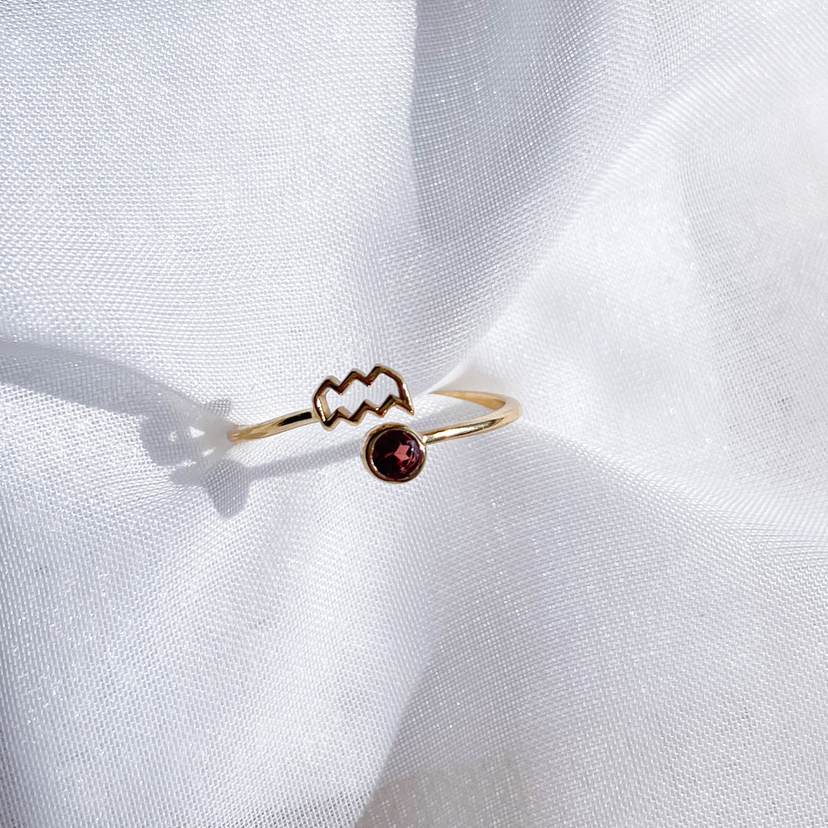 The Healing Hedge Witch’s Zodiac Gold Plated Ring, featuring a zigzag design with a small round red birthstone, is shown on textured white fabric—a stylish, adjustable piece for jewelry lovers.