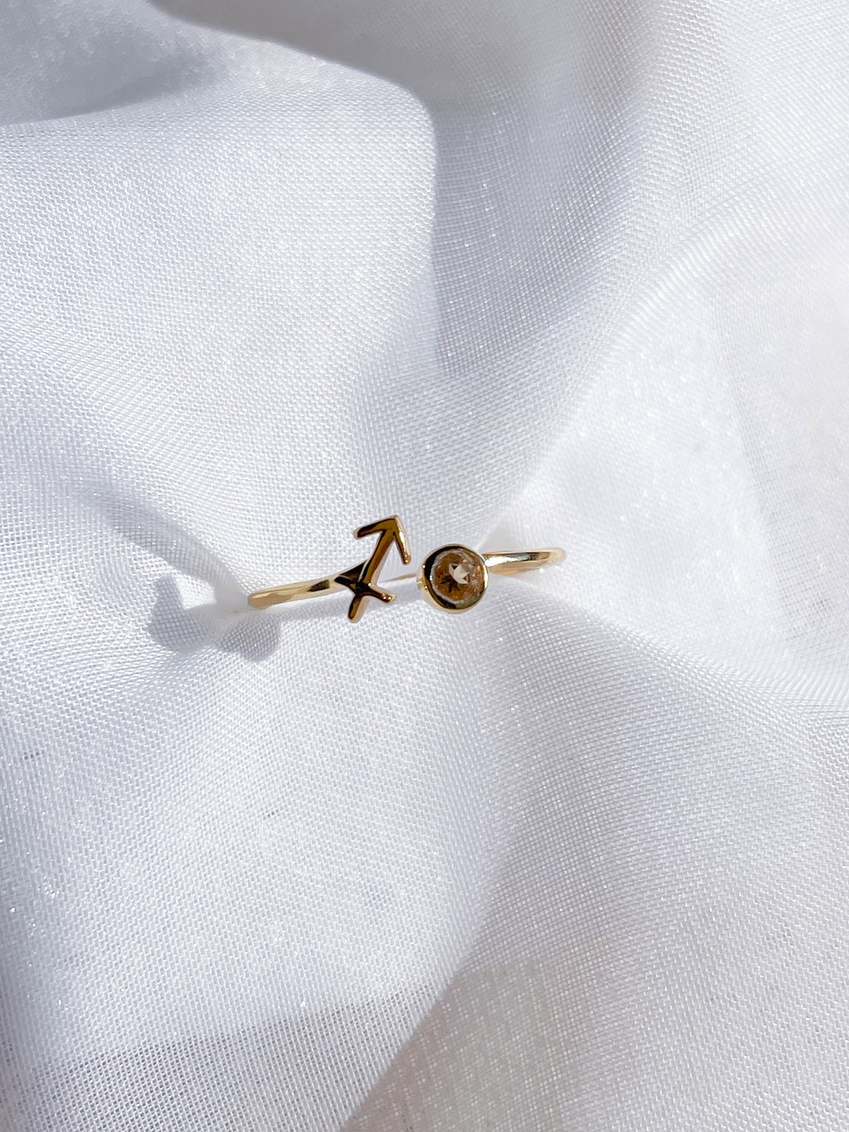 The Healing Hedge Witch's Zodiac Gold Plated Ring showcases a Sagittarius arrow on one end and a sparkling birthstone on the other—an elegant, adjustable piece displayed on soft white fabric.