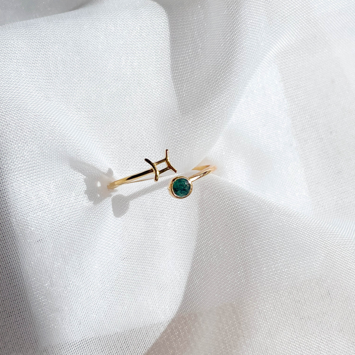 The Healing Hedge Witch's Zodiac Gold Plated Ring, featuring a Pisces symbol and a small green gemstone, is displayed on white textured fabric under soft lighting.