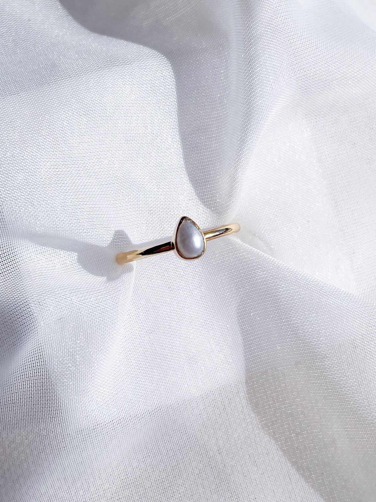 The Healing Hedge Witch's Pearl Stackable Crystal Ring Gold Plated features a teardrop-shaped pearl bezel set at the center, elegantly displayed on soft, white, translucent fabric with gentle folds.