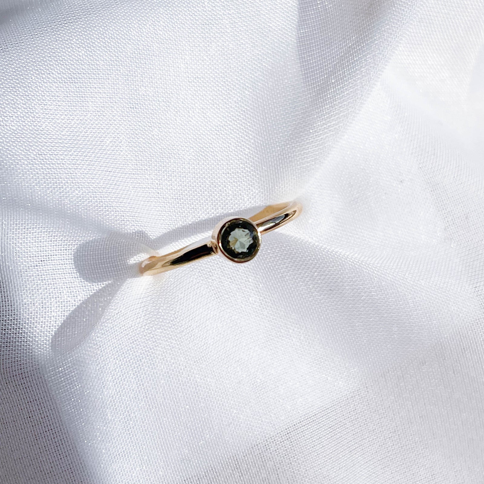 The Healing Hedge Witch's Moldavite Faceted Stackable Ring Gold Plated, featuring a single 3x5 mm round green moldavite gemstone in a delicate gold bezel setting, is displayed on softly wrinkled white fabric in bright natural light.