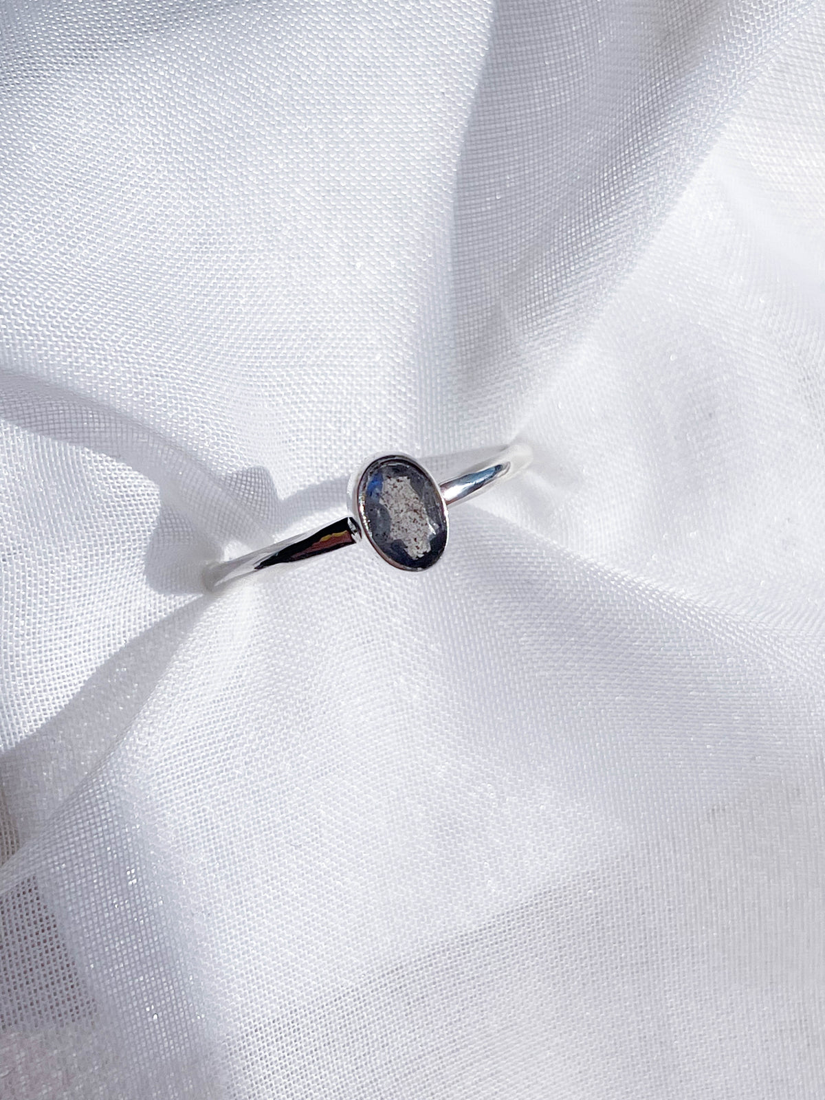 The Healing Hedge Witch’s Labradorite Faceted Stackable Ring, crafted from 925 sterling silver with a bezel-set, oval labradorite gemstone, rests on soft white fabric with gentle folds.