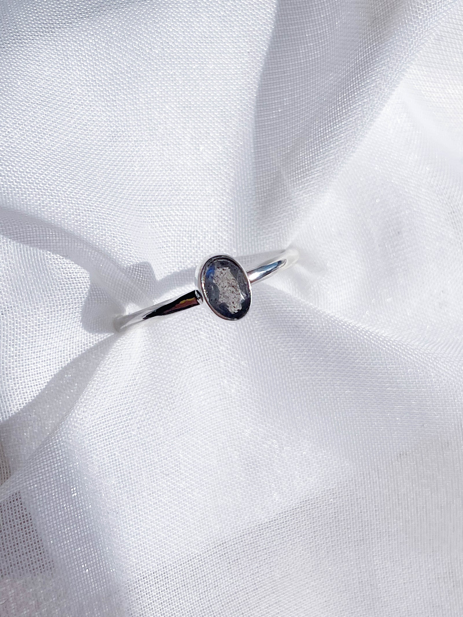 The Healing Hedge Witch’s Labradorite Faceted Stackable Ring, crafted from 925 sterling silver with a bezel-set, oval labradorite gemstone, rests on soft white fabric with gentle folds.