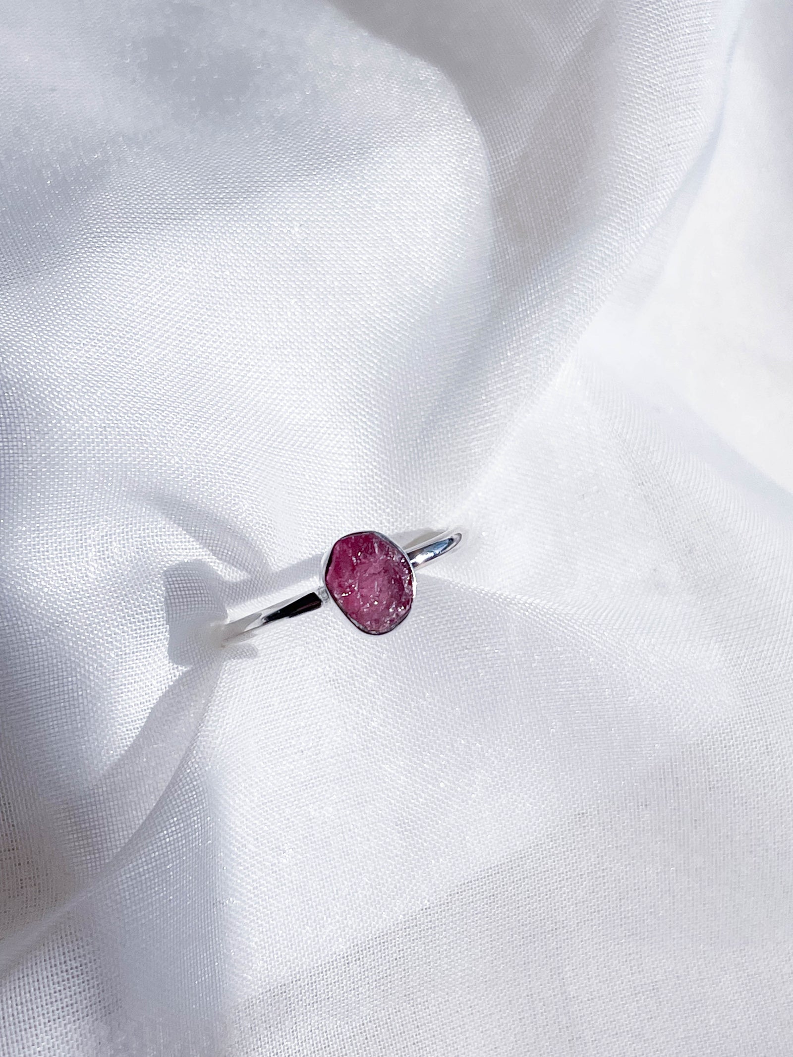 The Healing Hedge Witch's Pink Tourmaline Stackable Rough Crystal Ring features a simple, elegant silver band with a round pink stone centerpiece, displayed on a soft white textured fabric background.