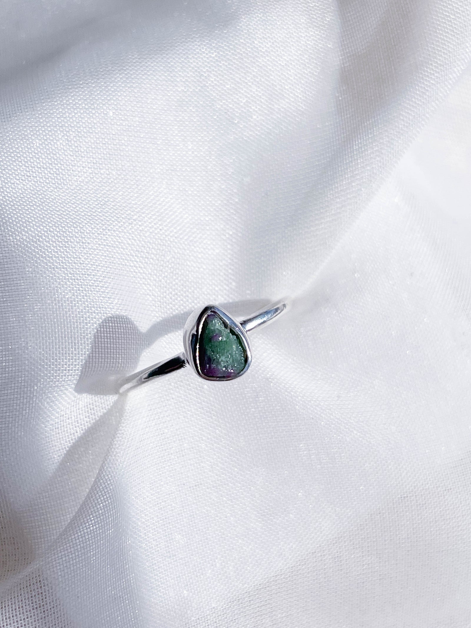 The Healing Hedge Witch's Ruby Zoisite Rough Crystal Stackable Ring, featuring a small, irregularly shaped green and purple stone, is displayed on soft, white textured fabric.