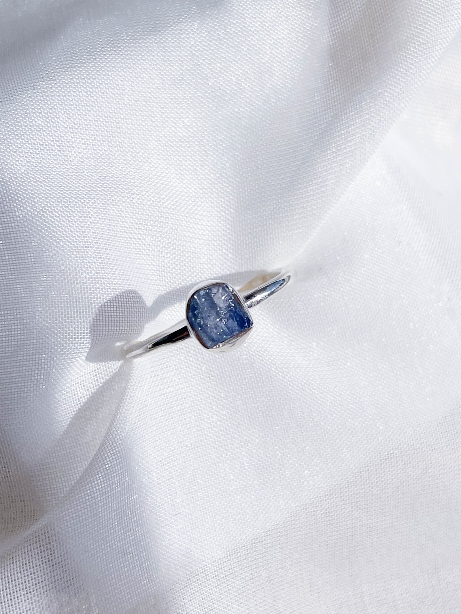 The Healing Hedge Witch Blue Sapphire Stackable Rough Crystal Ring is shown on a white textured fabric background in bright lighting.