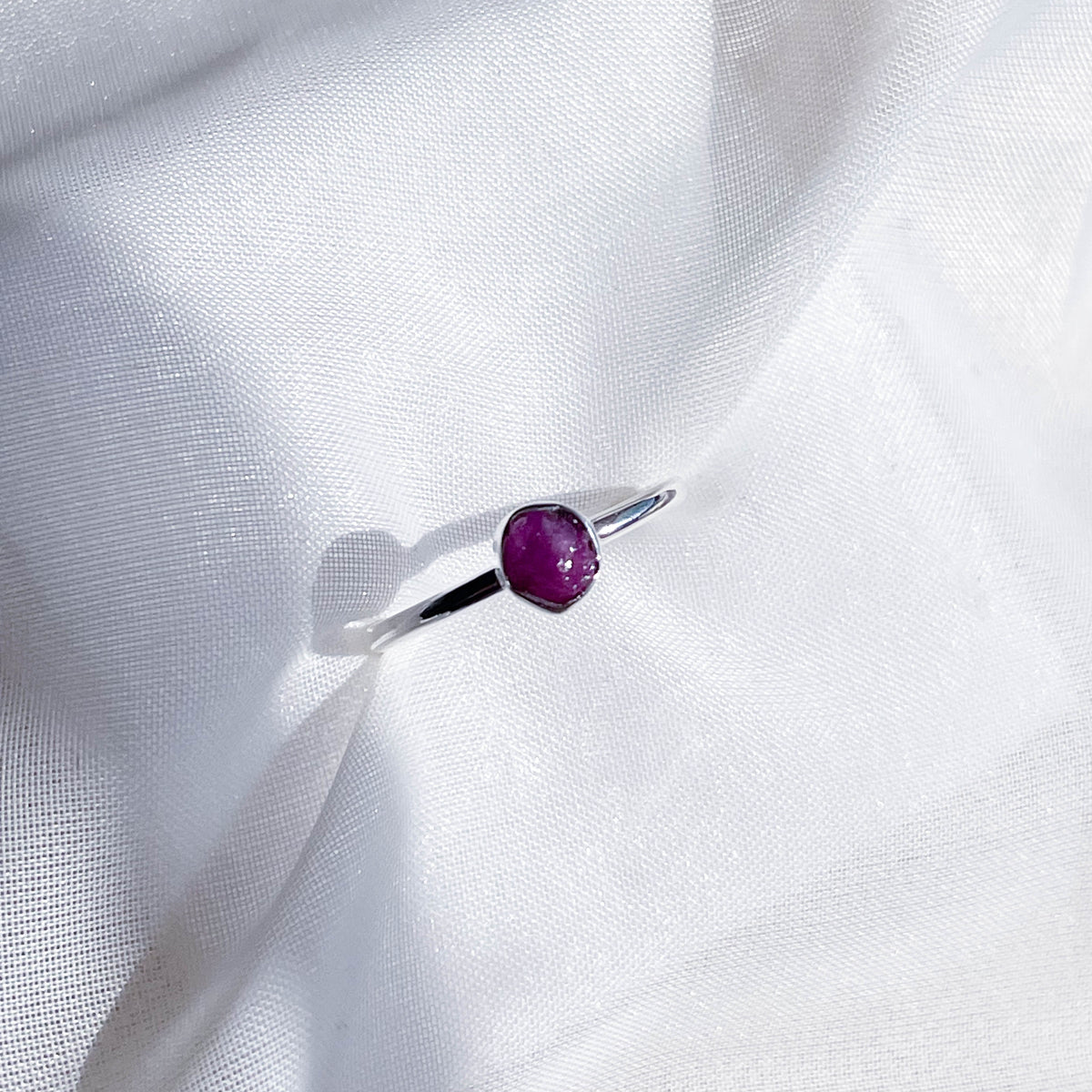 The Healing Hedge Witch's Ruby Stackable Rough Crystal Ring is shown on white textured fabric under bright light, featuring a small, irregular ruby gemstone set in a silver band.