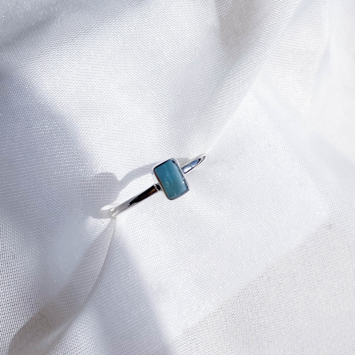 The Healing Hedge Witch's Larimar Stackable Rough Crystal Ring in 925 Sterling Silver, featuring a small blue rectangular stone, rests on soft white sheer fabric under natural light—ideal for throat chakra balance.
