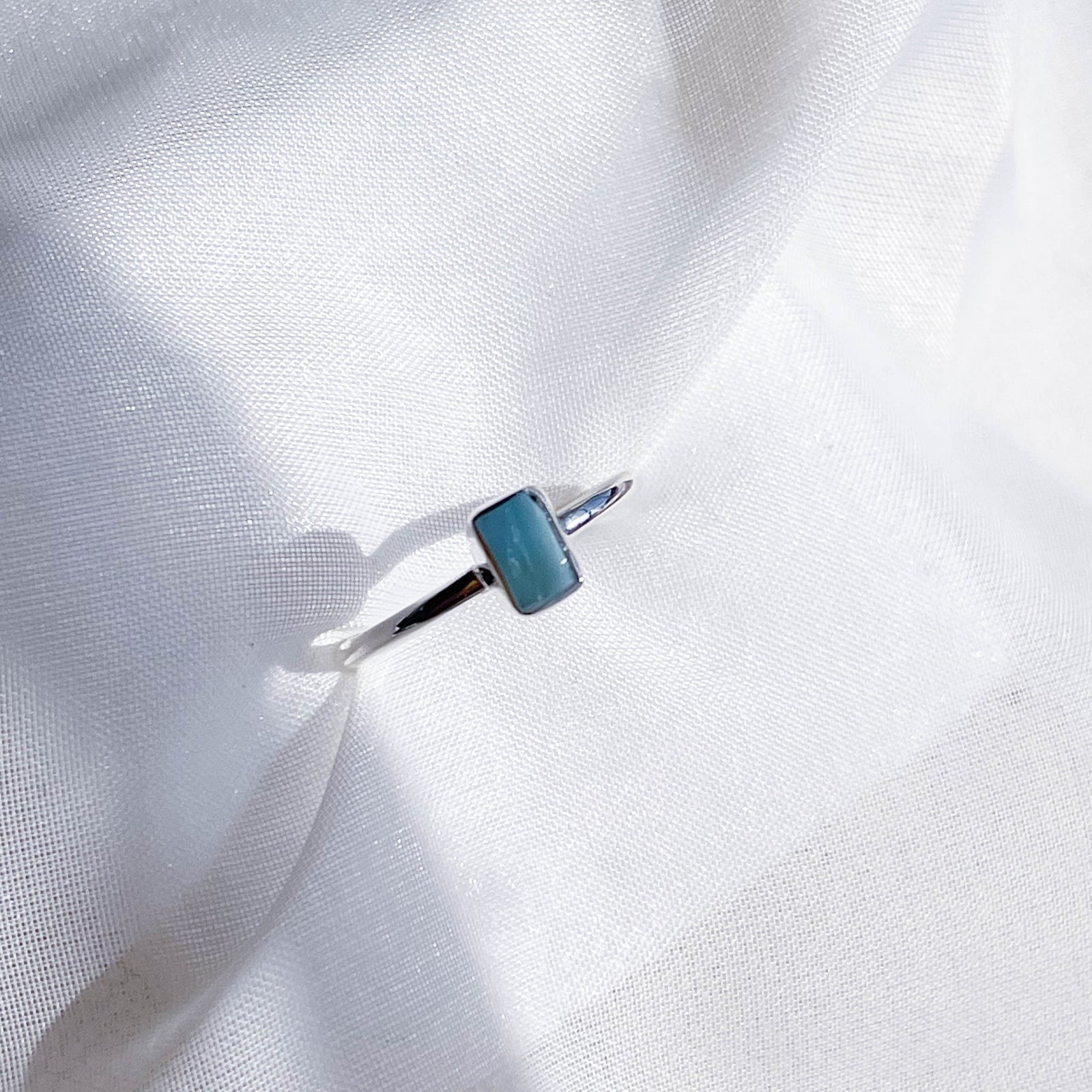 The Healing Hedge Witch's Larimar Stackable Rough Crystal Ring in 925 Sterling Silver, featuring a small blue rectangular stone, rests on soft white sheer fabric under natural light—ideal for throat chakra balance.