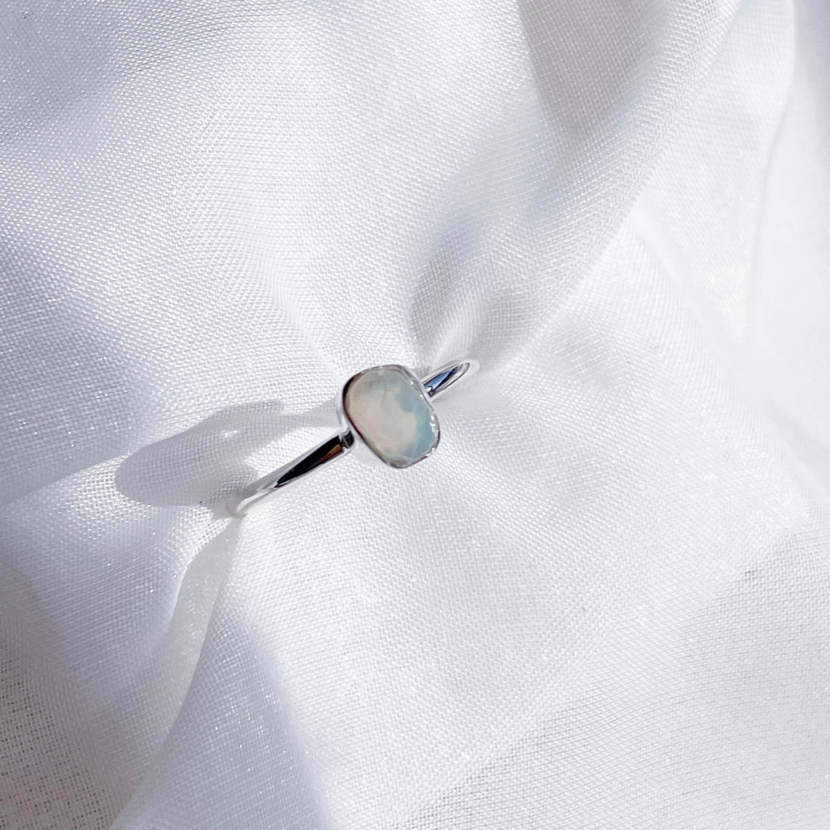 The Healing Hedge Witch's Ethiopian Opal Stackable Rough Crystal Ring, featuring an oval, translucent pale blue opal, is displayed on softly draped white fabric in bright natural light.