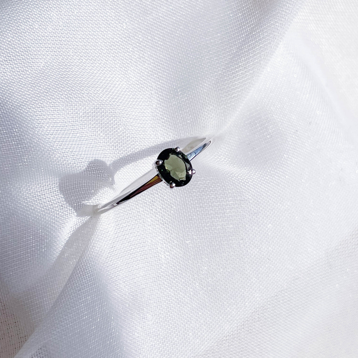 The Healing Hedge Witch Moldavite Faceted Stackable Ring features a delicate 925 sterling silver band with a 3x5 mm oval green gemstone in four prongs, shown on white textured fabric under bright lighting.