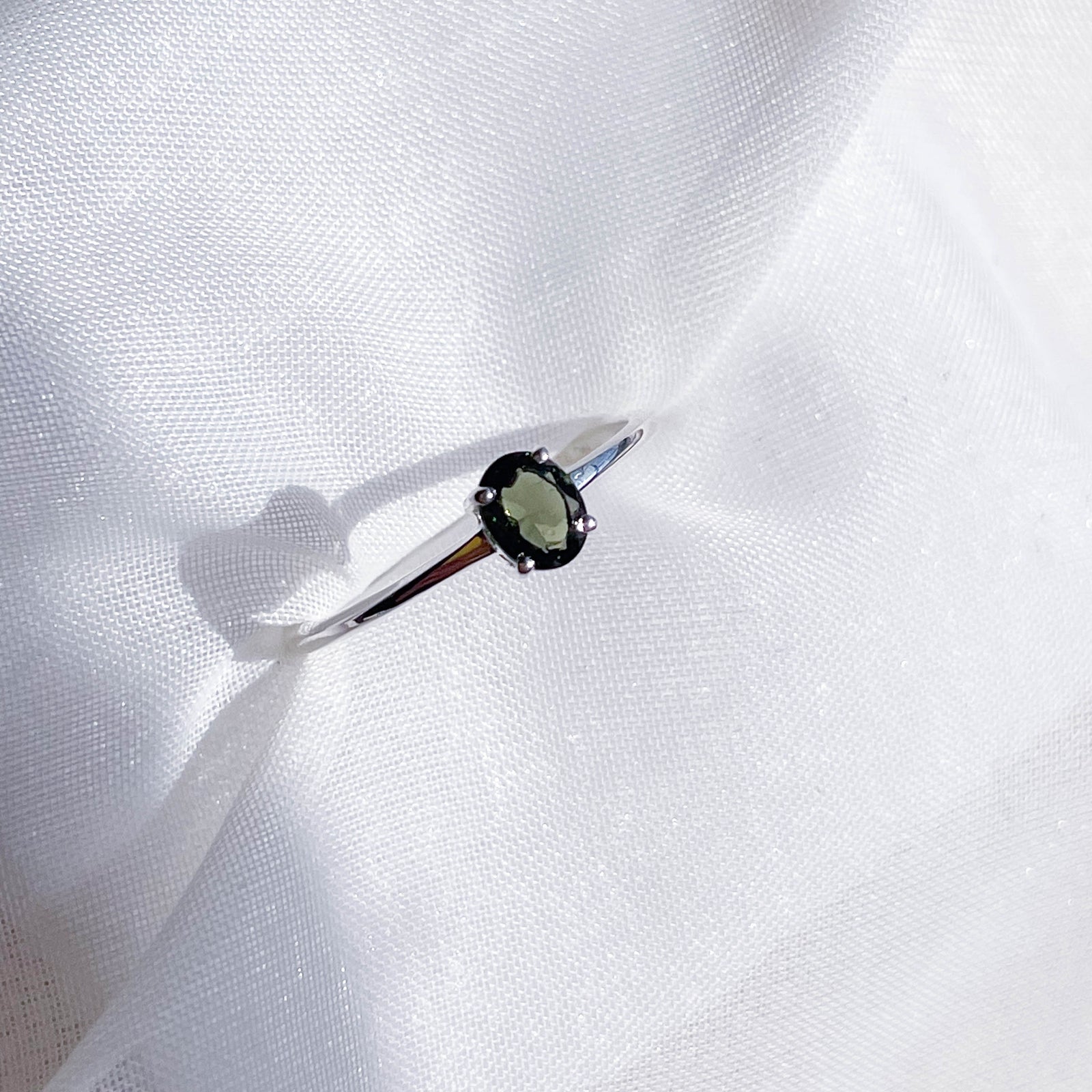 The Healing Hedge Witch Moldavite Faceted Stackable Ring features a delicate 925 sterling silver band with a 3x5 mm oval green gemstone in four prongs, shown on white textured fabric under bright lighting.