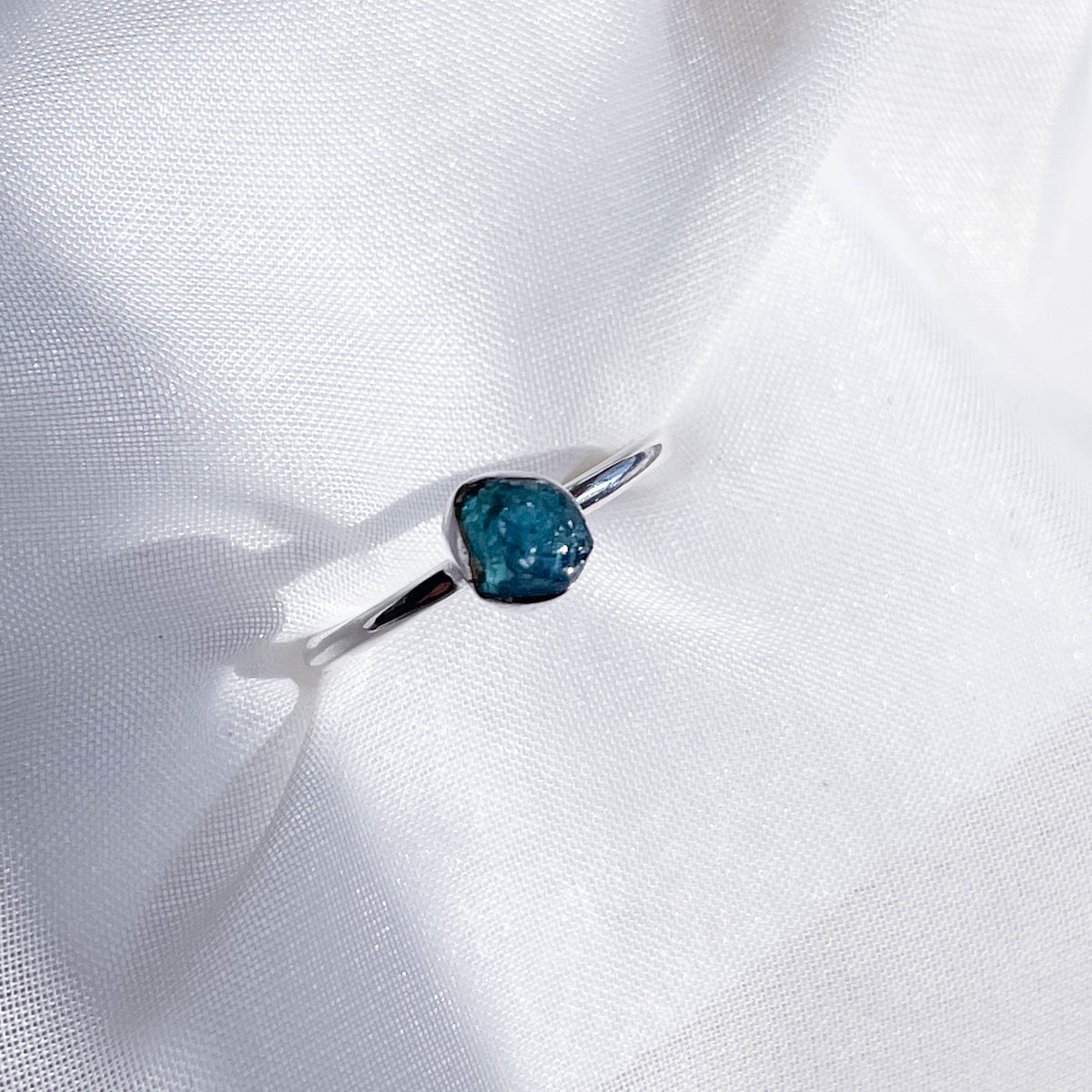 The Healing Hedge Witch's Blue Tourmaline Stackable Rough Crystal Ring, featuring an oval blue gemstone, is displayed on softly textured white fabric under natural light.